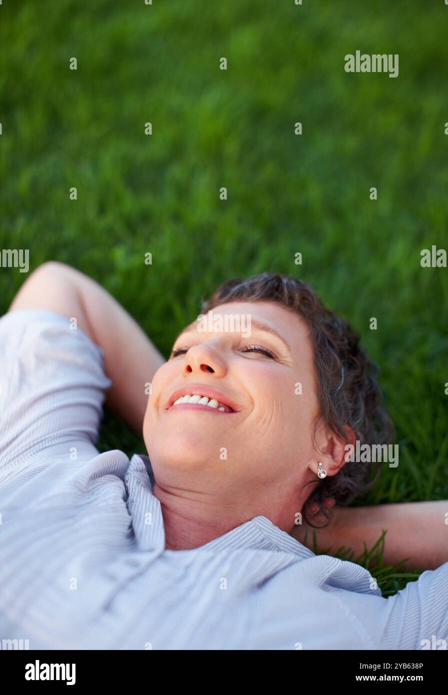 Donna, sorridi e rilassati sullo spazio erboso per il comfort, la pace e la salute mentale positiva. Calma, persona matura e felice nel parco naturale per i viaggi estivi Foto Stock