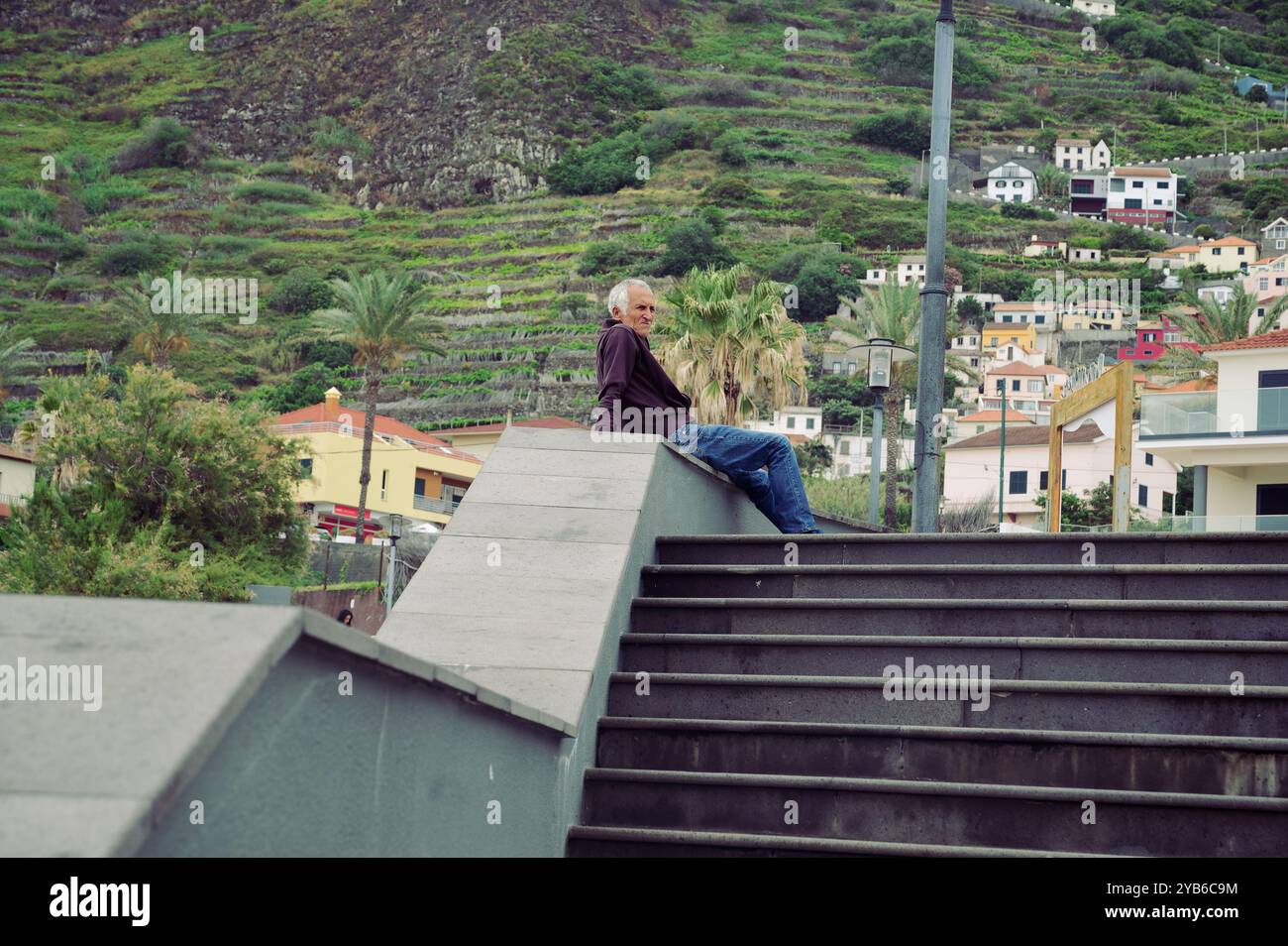 Un uomo anziano si rilassa sulle scale, affacciato sulle colline terrazzate e sulle case colorate in un tranquillo villaggio di Madeira Foto Stock