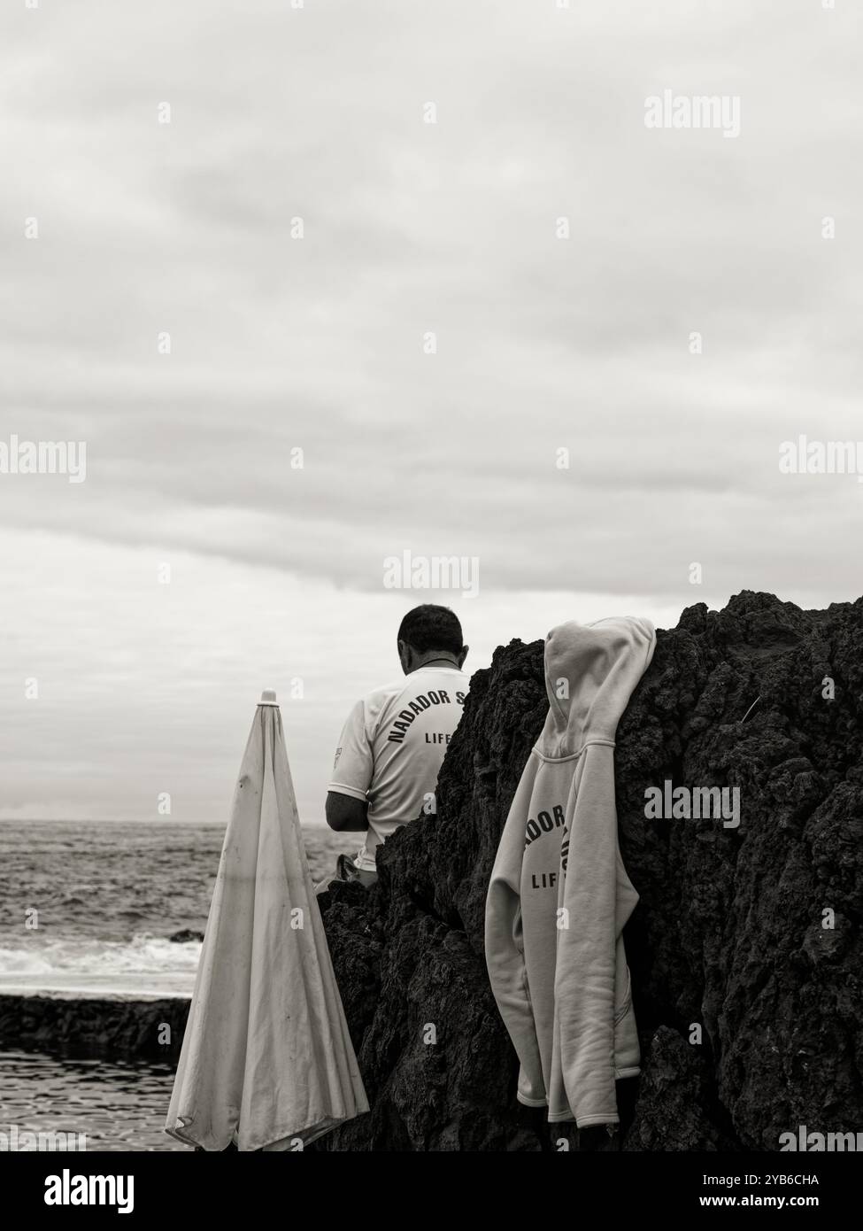 Monochorome, bagnino si erge tra rocce vulcaniche, con una felpa con cappuccio drappeggiata sulla pietra, che si affaccia sull'oceano presso le piscine naturali di Porto Moniz Foto Stock