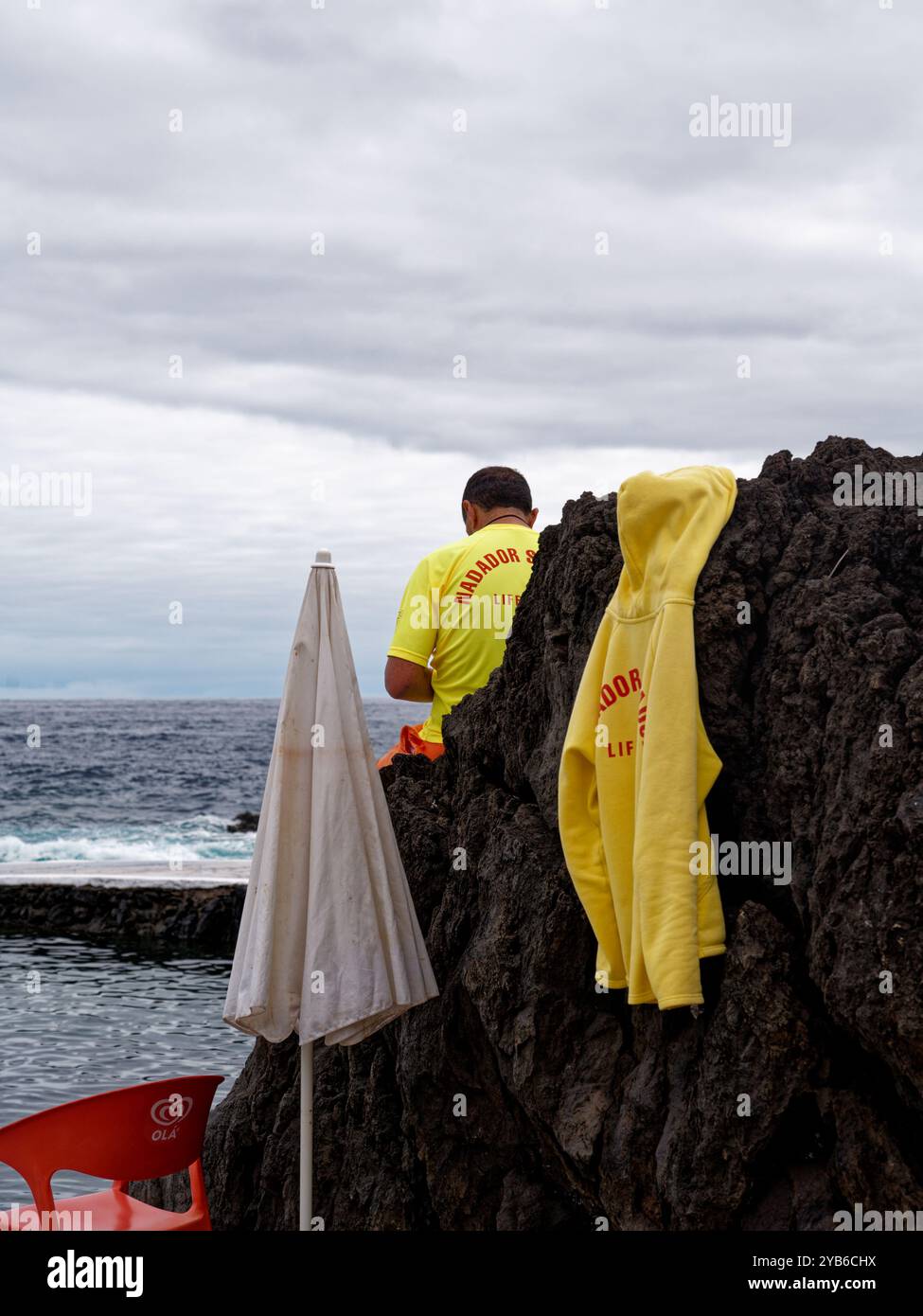 Un bagnino siede su un masso vulcanico, con una felpa gialla drappeggiata sulla pietra, che si affaccia sull'oceano presso le piscine naturali di Porto Moniz Foto Stock