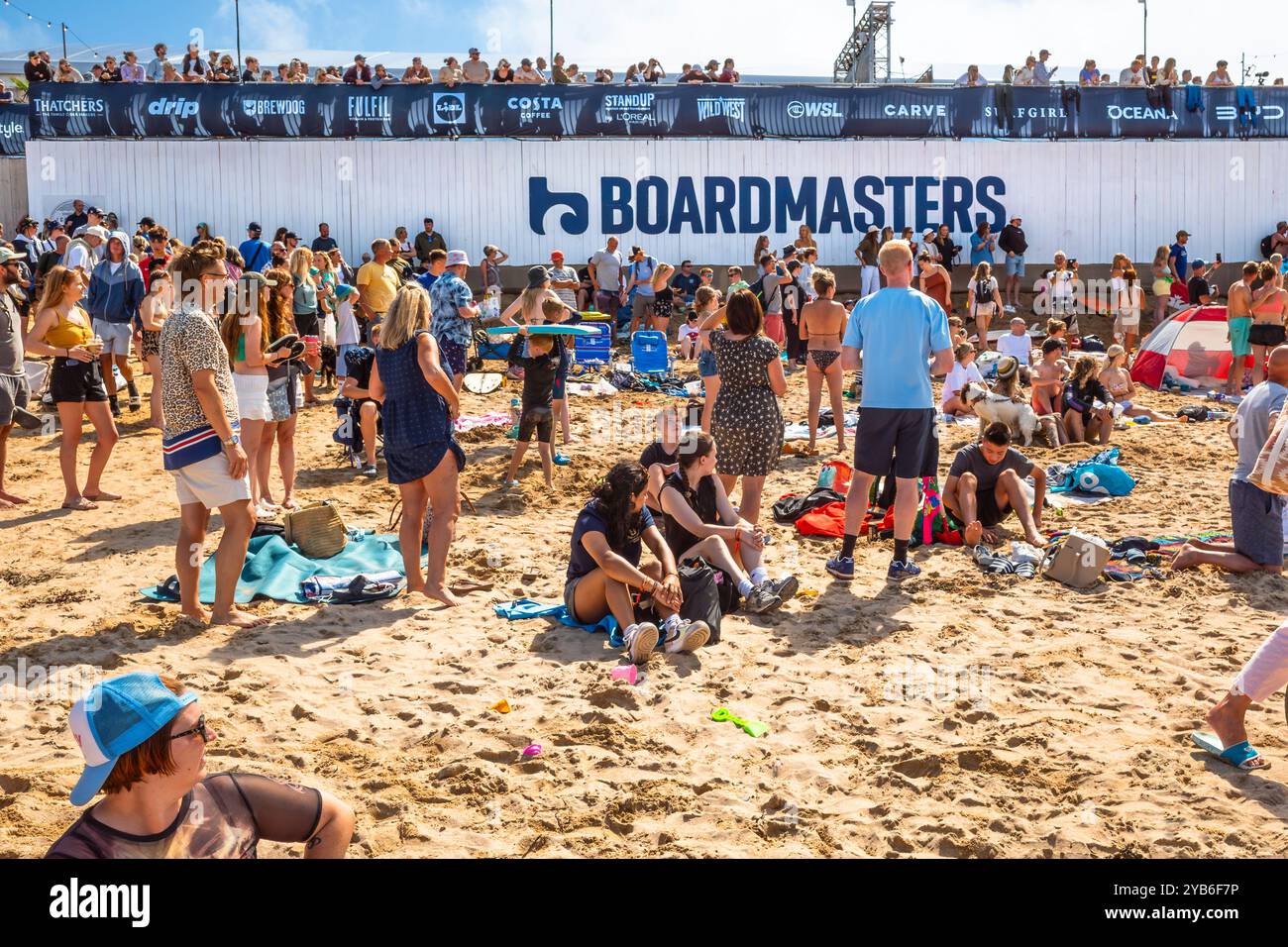 I vacanzieri gli spettatori che fanno surf si divertono al Boardmasters Surfing Festival, godendosi il sole estivo sull'iconica spiaggia di Fistral, famosa in tutto il mondo Foto Stock