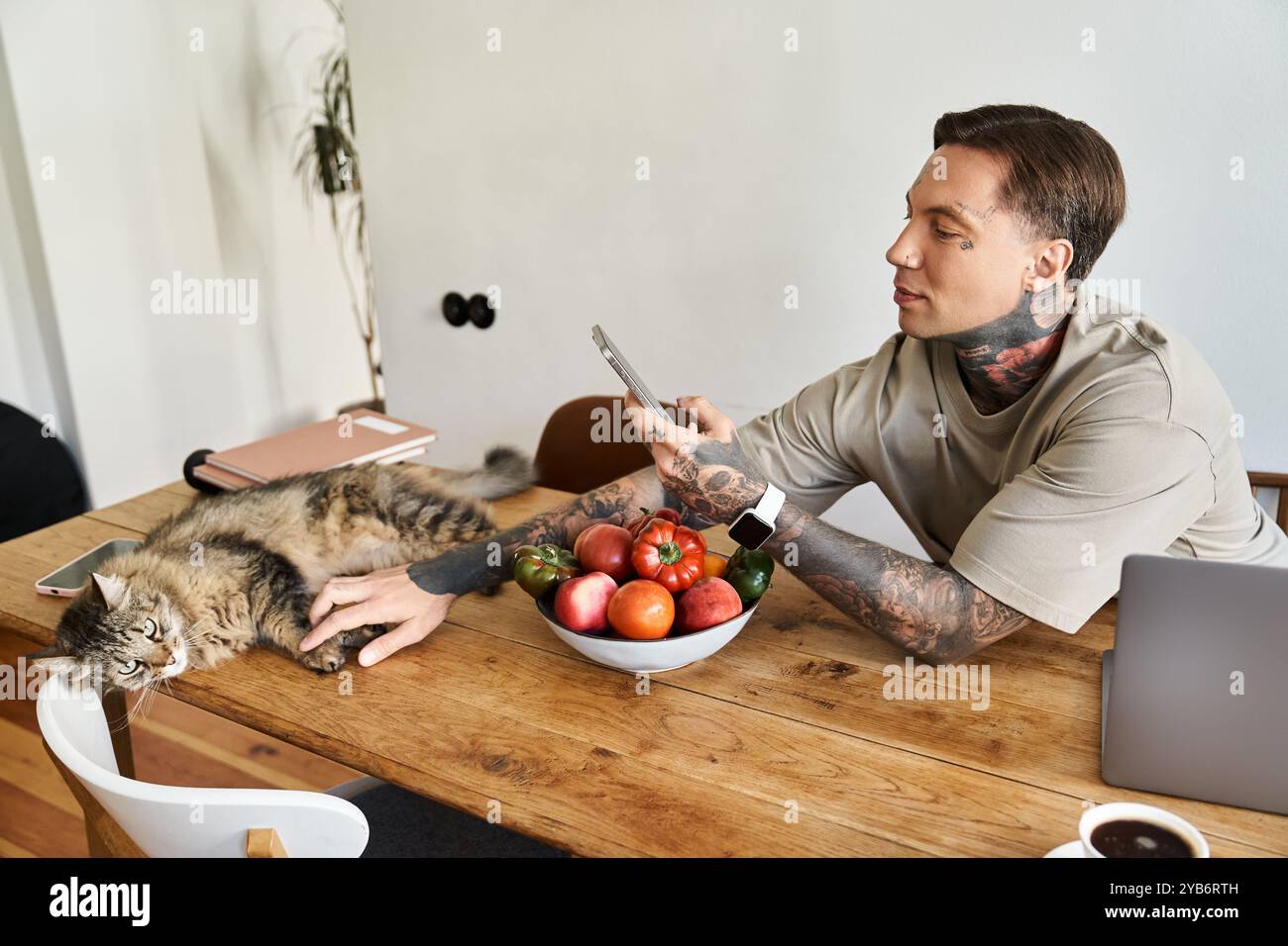 Un giovane uomo si impegna con il suo telefono mentre si rilassa a casa accanto al suo gatto rilassato e ai suoi prodotti freschi. Foto Stock