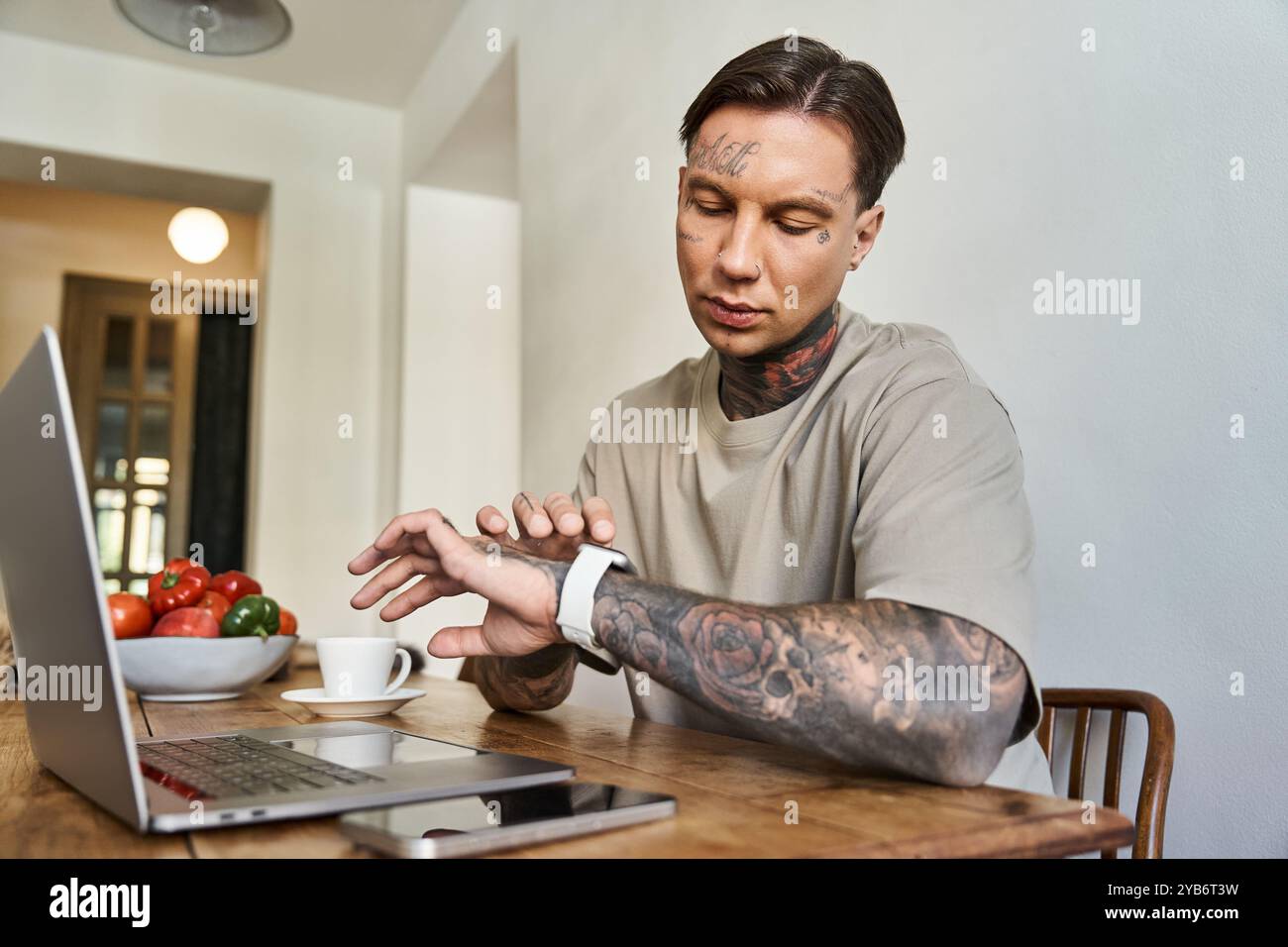 Un giovane uomo tatuato si concentra intensamente sul suo notebook mentre guarda il suo smartwatch. Foto Stock