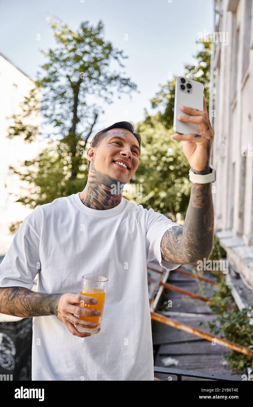 Un giovane con i tatuaggi sorride in grande, catturando un momento con un drink in mano sul suo balcone. Foto Stock
