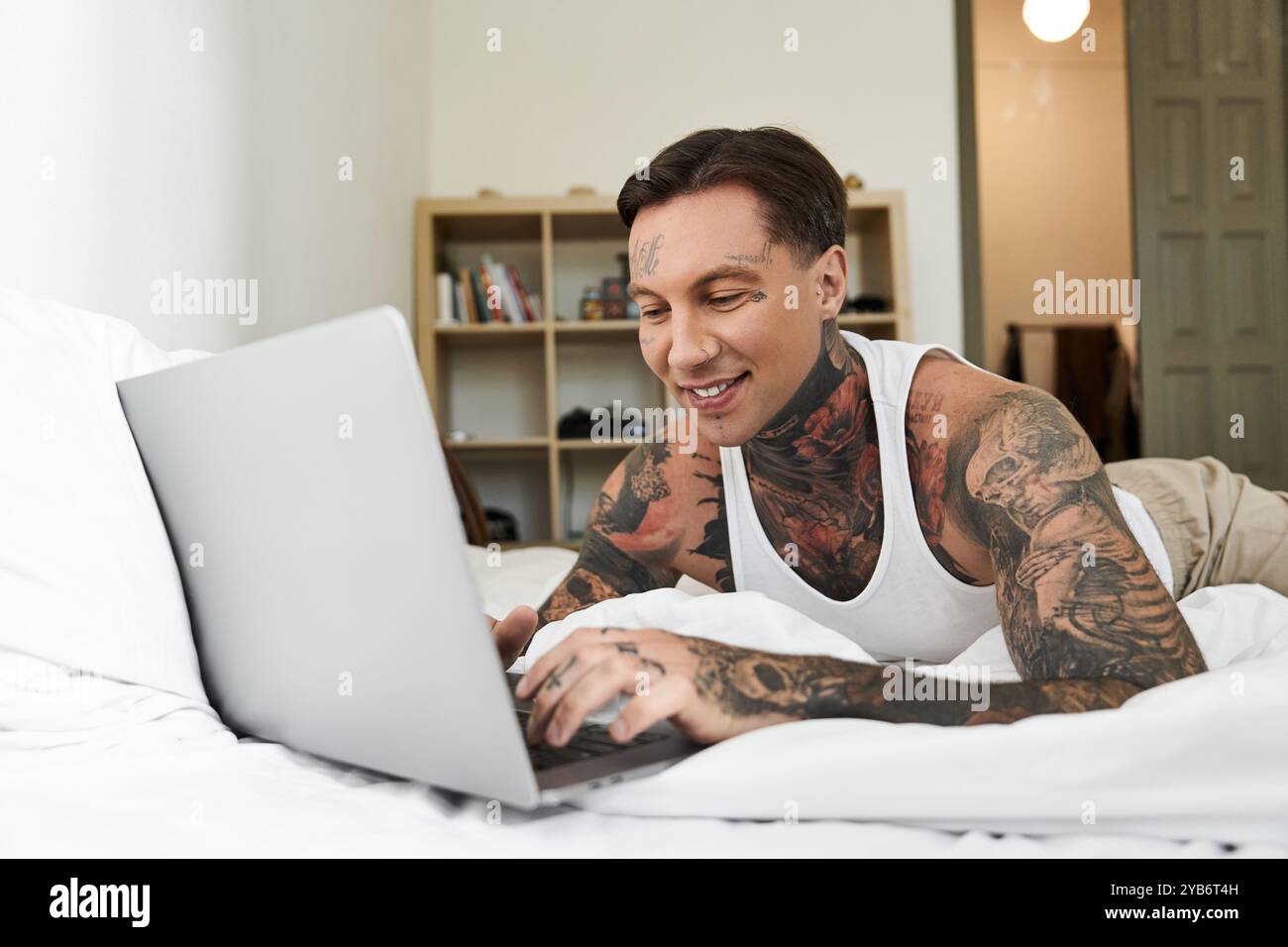 Un giovane e bello uomo si rilassa sul letto, si affida al suo portatile, sorridendo felicemente. Foto Stock