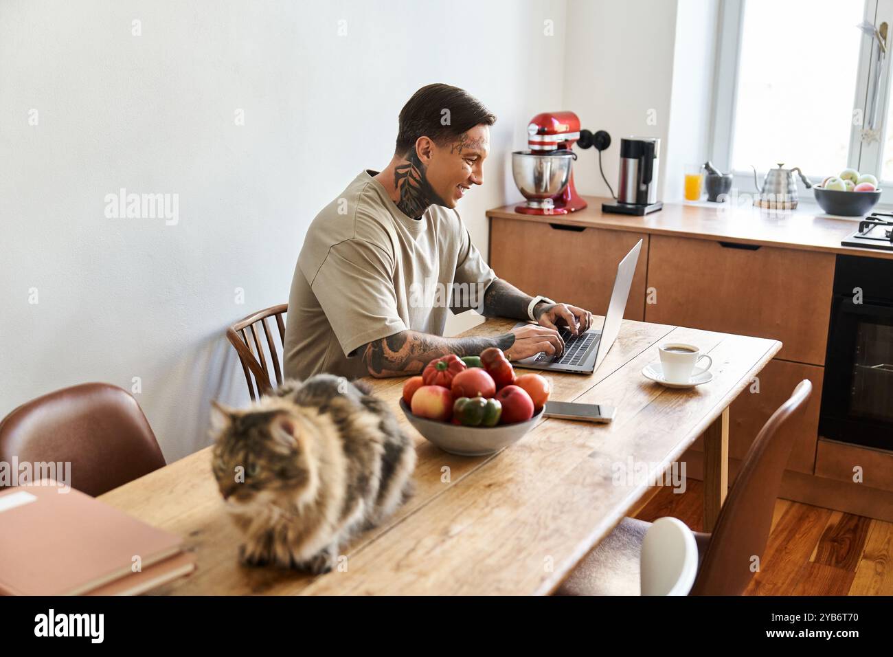 Il giovane si occupa di un lavoro concentrato sul suo portatile, circondato da frutta fresca e un gatto curioso. Foto Stock