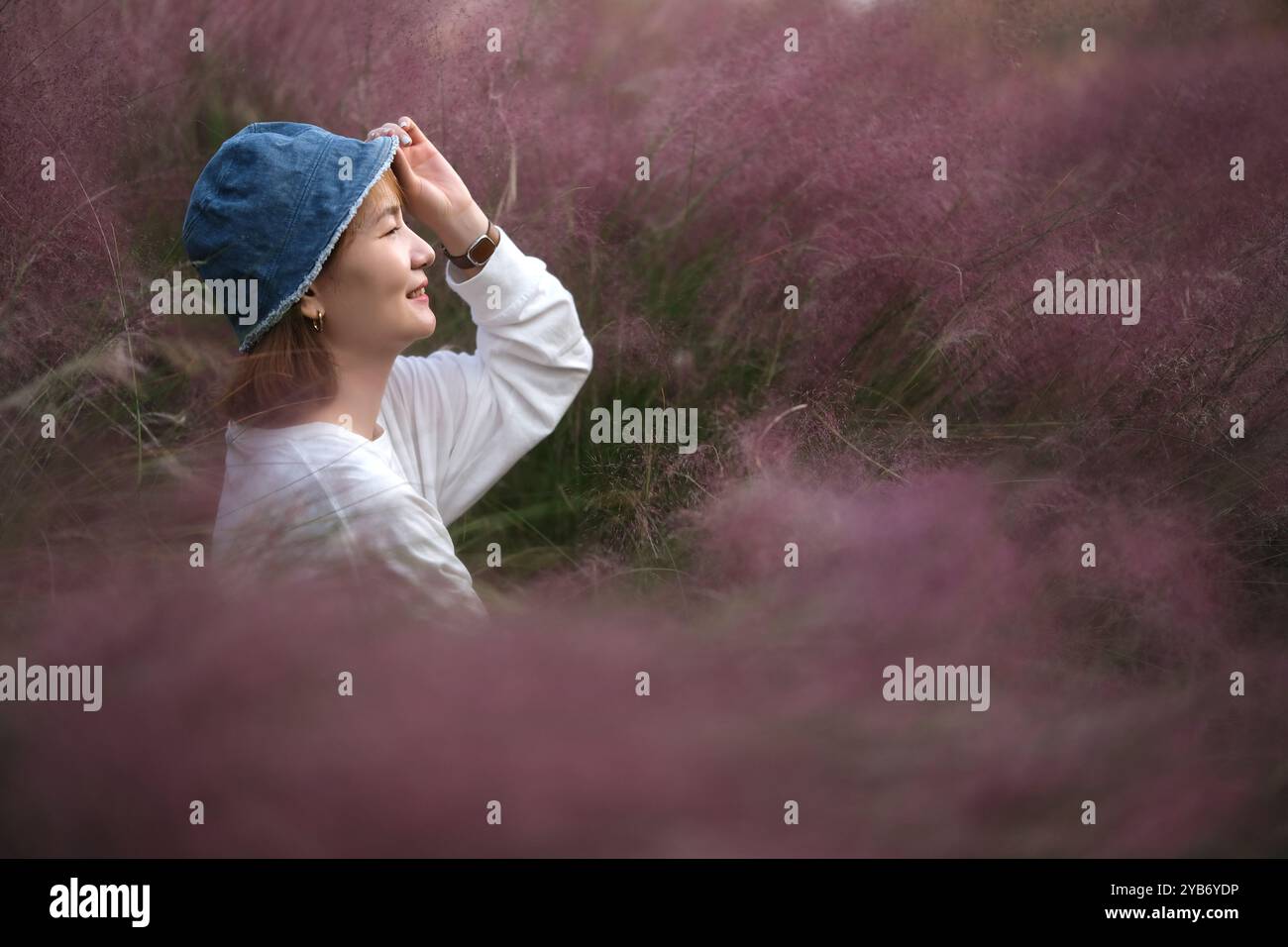 Sorridi giovane donna asiatica con cappello in mano, vista laterale. circondato da erba di muhly (muhlenbergia capillaris) viola Foto Stock