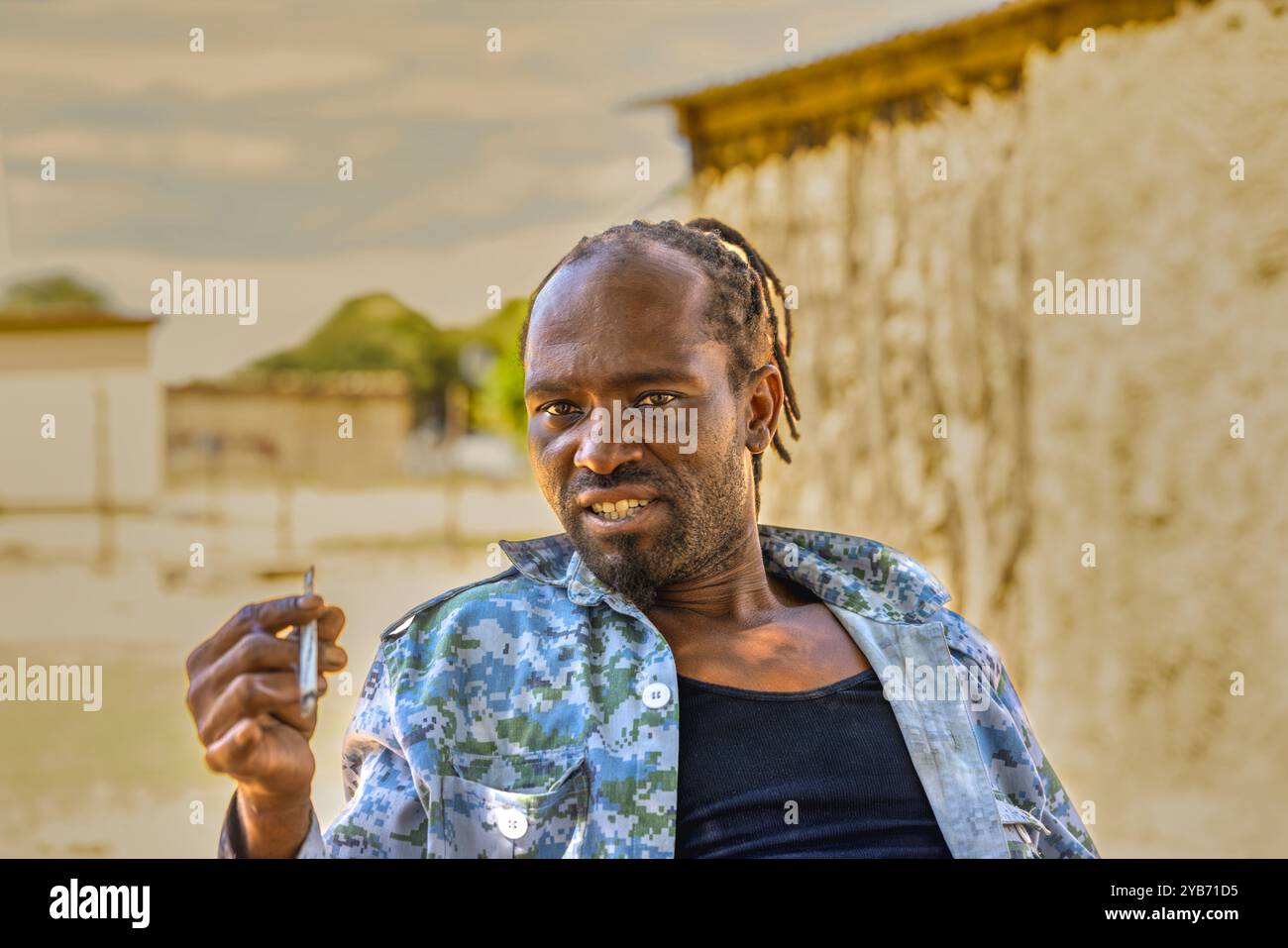 uomo africano del villaggio con dreadlock in piedi davanti alla casa che fuma marijuana Foto Stock
