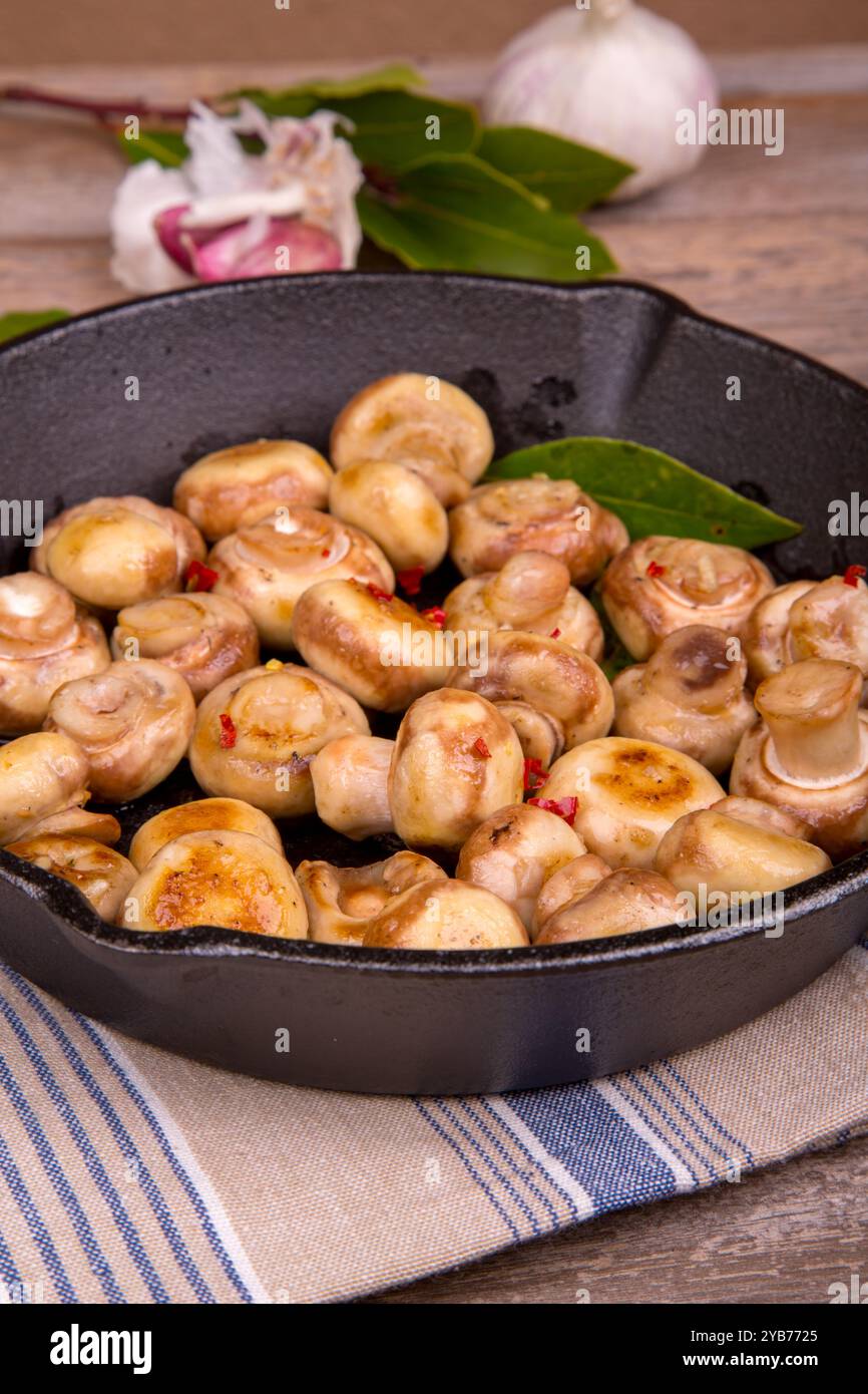 Una padella in ghisa piena di funghi saltati. I funghi sono cotti alla perfezione con fiocchi di peperoncino, aglio ed erbe aromatiche. Un telo da tè e rustico Foto Stock