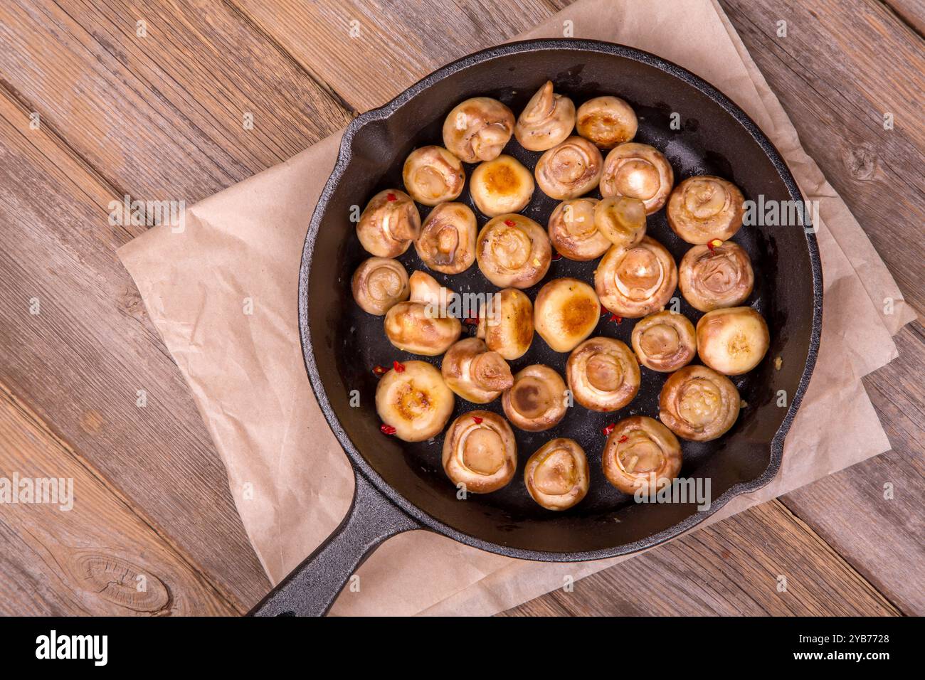 Una padella in ghisa piena di funghi saltati. I funghi sono cotti alla perfezione con i fiocchi di peperoncino. Carta resistente al grasso e bac in legno rustico Foto Stock
