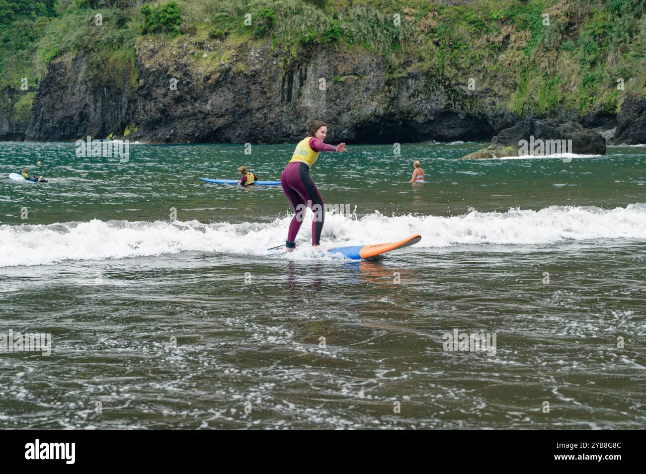 Una giovane surfista perfeziona il suo equilibrio su una piccola onda durante una lezione di surf alla spiaggia di Seixal, con rigogliose scogliere sullo sfondo Foto Stock