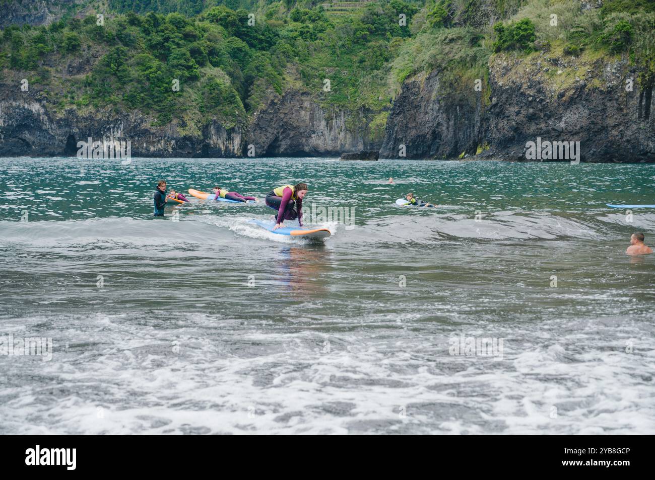Una giovane surfista pratica in piedi sulla sua tavola durante una lezione di surf alla spiaggia di Seixal, con altri studenti e rigogliose scogliere sullo sfondo Foto Stock