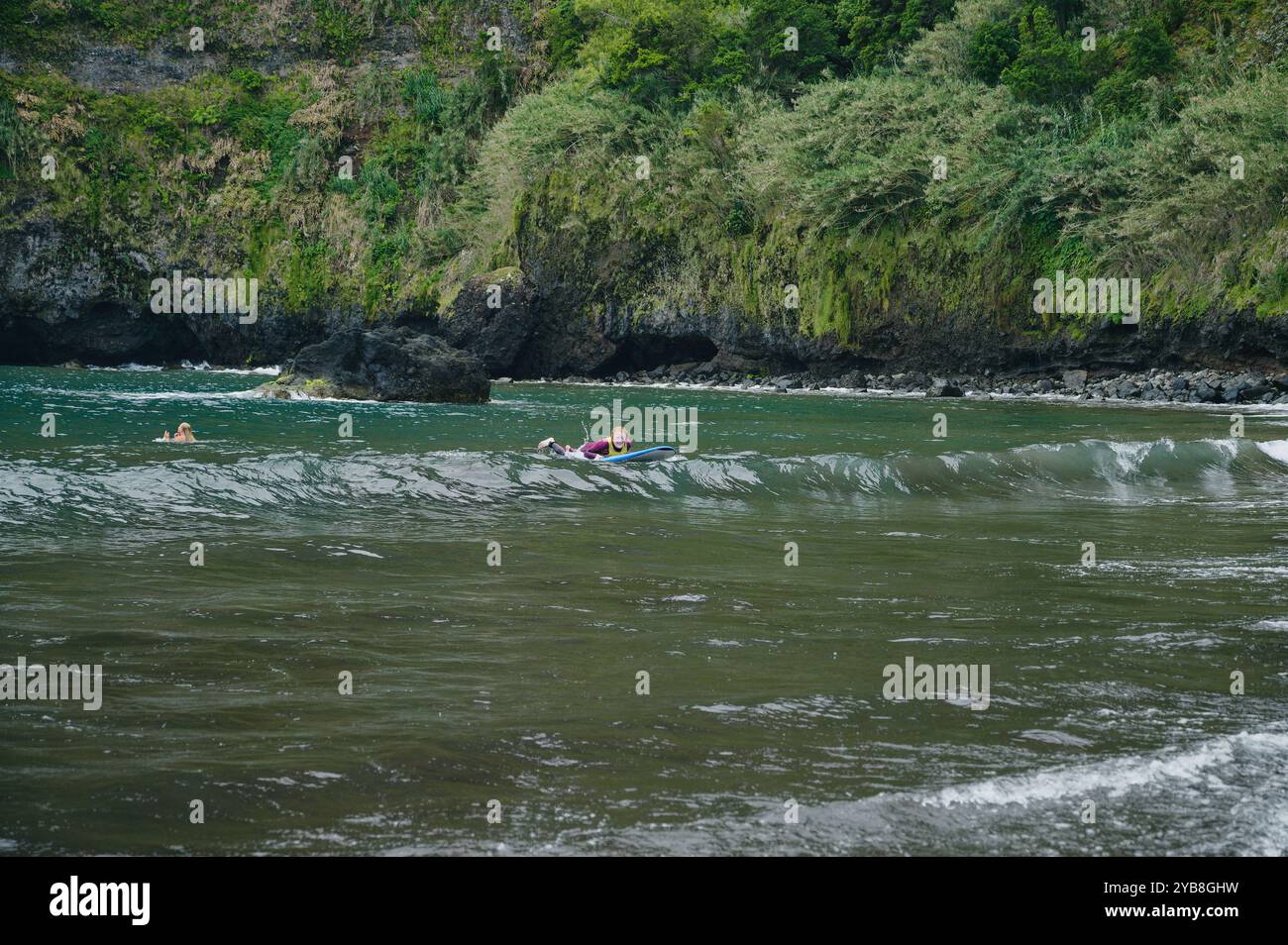 Un giovane surfista pagaia durante una lezione di surf alla spiaggia di Seixal, con rigogliose scogliere sullo sfondo Foto Stock