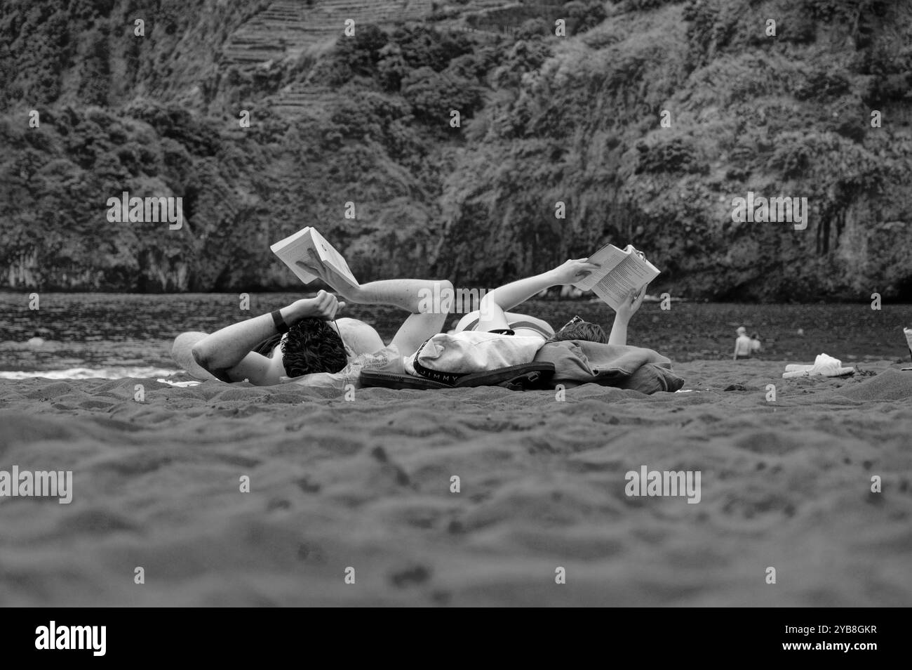 Una coppia si legge fianco a fianco sulla sabbia nera della spiaggia di Seixal, Madeira, con lussureggianti scogliere sullo sfondo (bianco e nero) Foto Stock