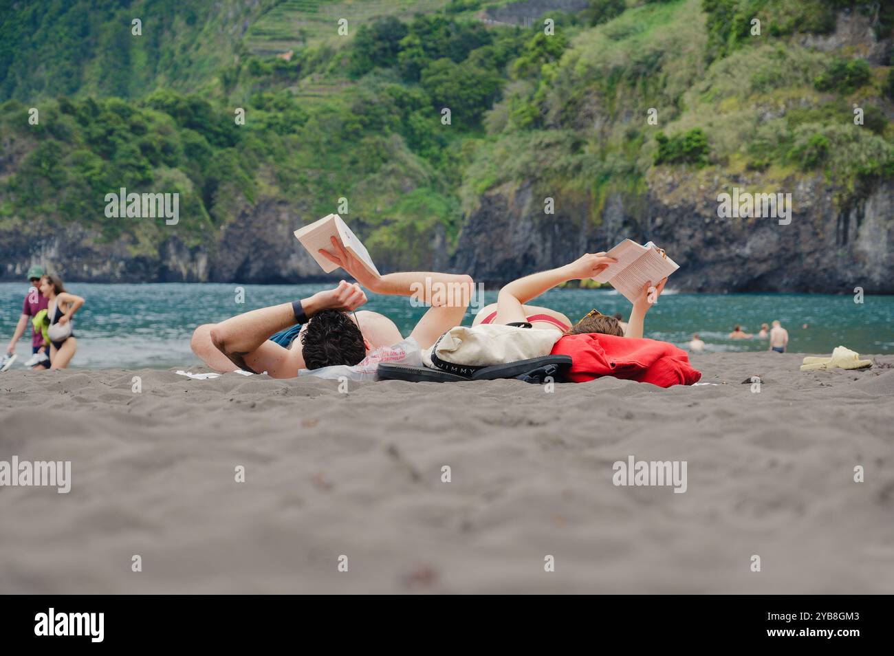 La coppia trascorre una giornata rilassante leggendo sulla sabbia nera della spiaggia di Seixal, con lussureggianti scogliere e acque turchesi a Madeira Foto Stock
