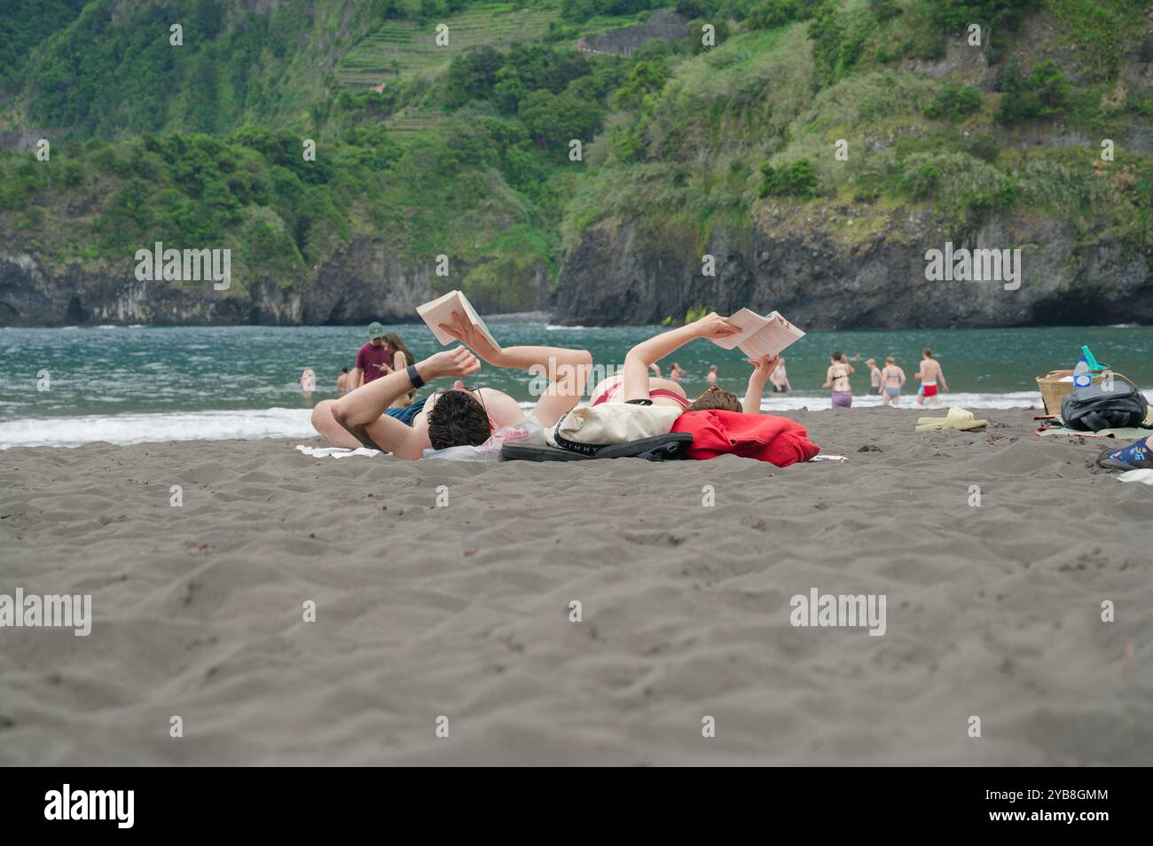 La coppia trascorre una giornata rilassante leggendo sulla sabbia nera della spiaggia di Seixal, con lussureggianti scogliere e acque turchesi a Madeira Foto Stock