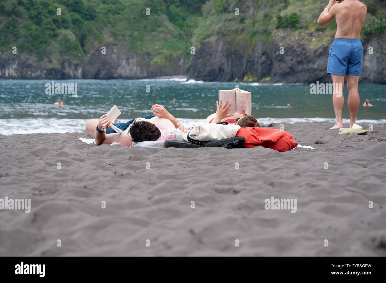 La coppia trascorre una giornata rilassante leggendo sulla sabbia nera della spiaggia di Seixal, con lussureggianti scogliere e acque turchesi a Madeira Foto Stock
