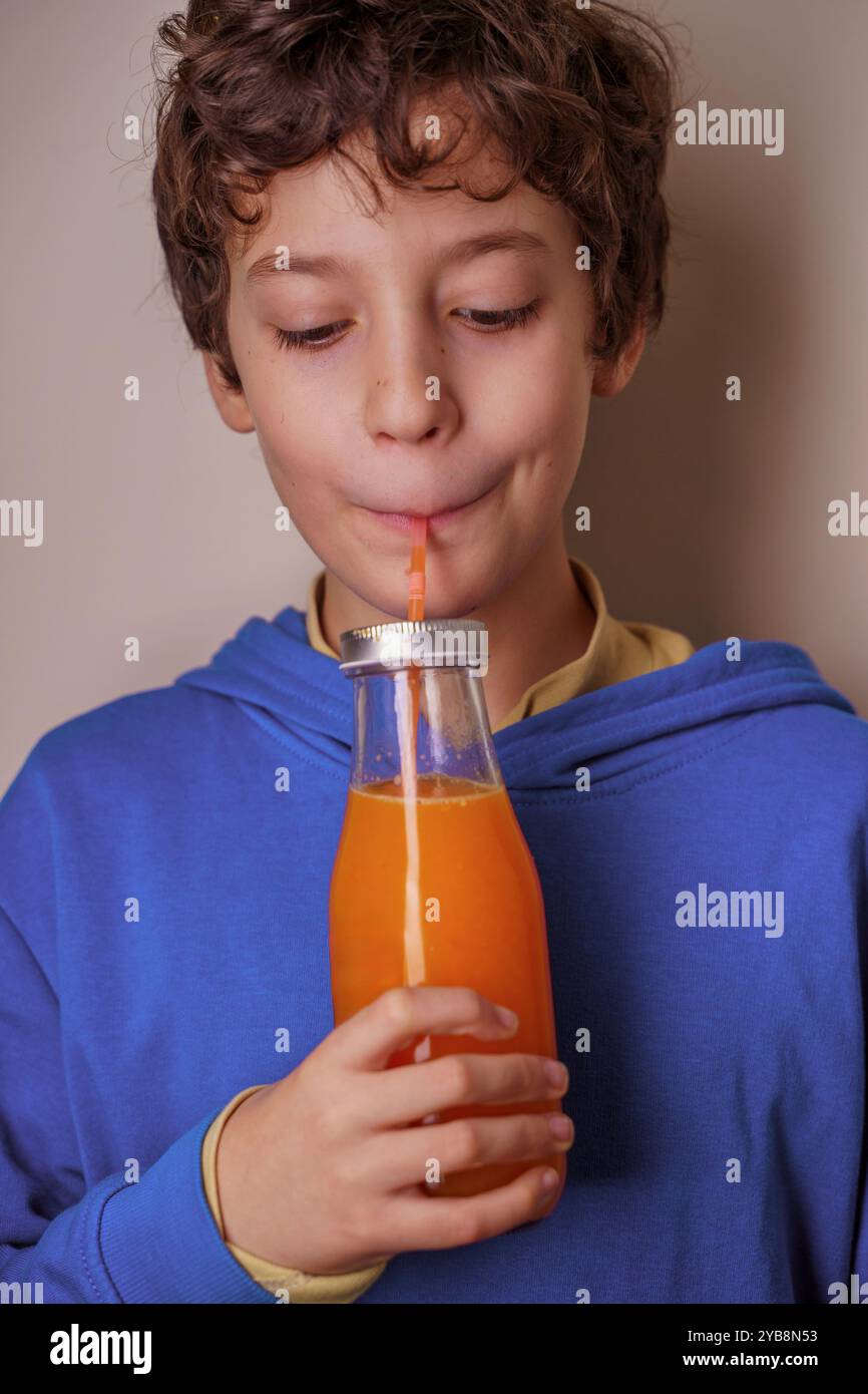 Ritratto di un ragazzo carino con capelli ricci sorseggiando succo d'arancia attraverso una cannuccia da una bottiglia di vetro. Il bambino indossa una felpa con cappuccio blu e dimostra una guarigione Foto Stock