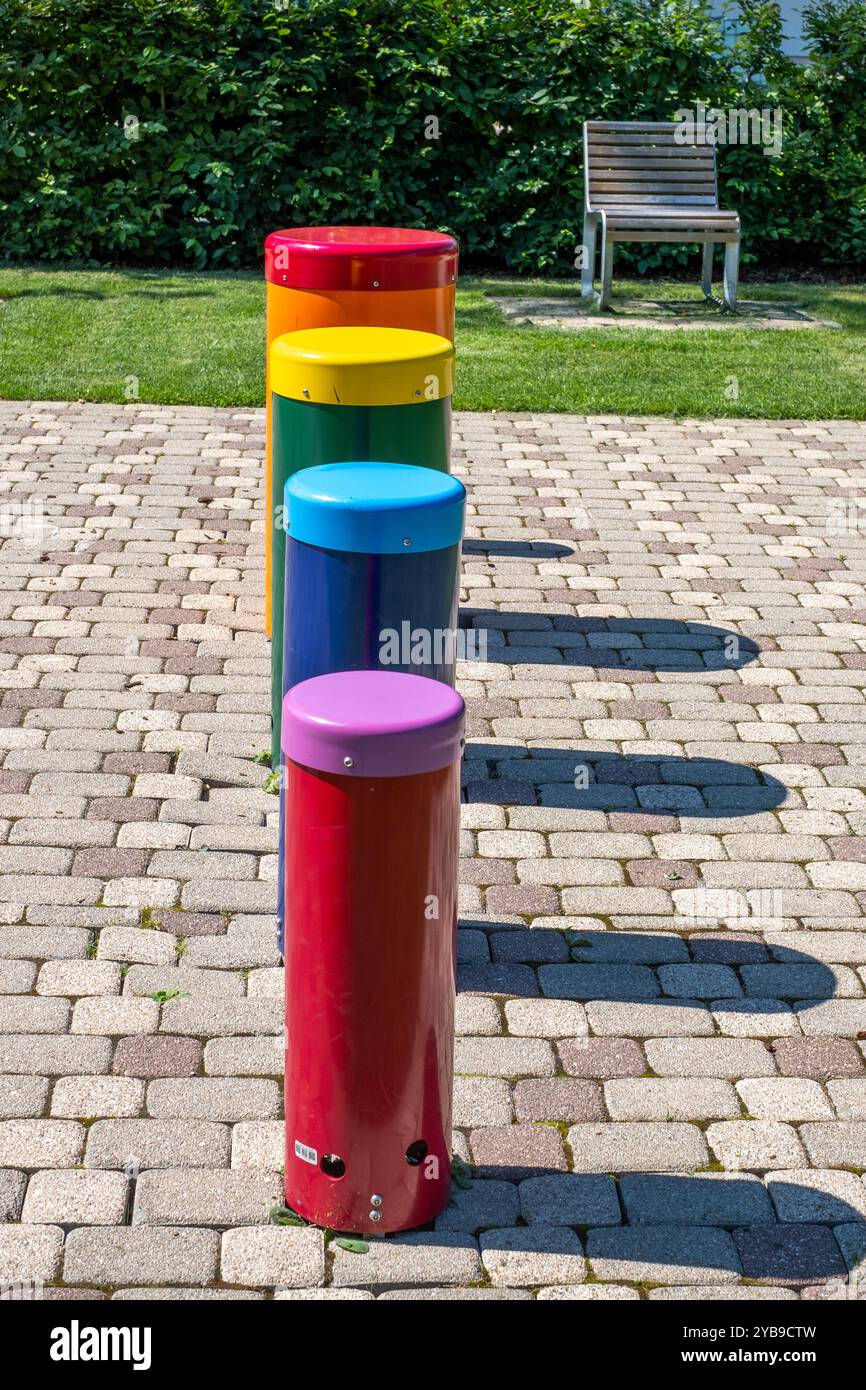 Una fila di vibranti strutture cilindriche in vari colori sorge su un sentiero lastricato accanto a una panchina del parco sotto cieli limpidi, creando un'atmosfera giocosa Foto Stock