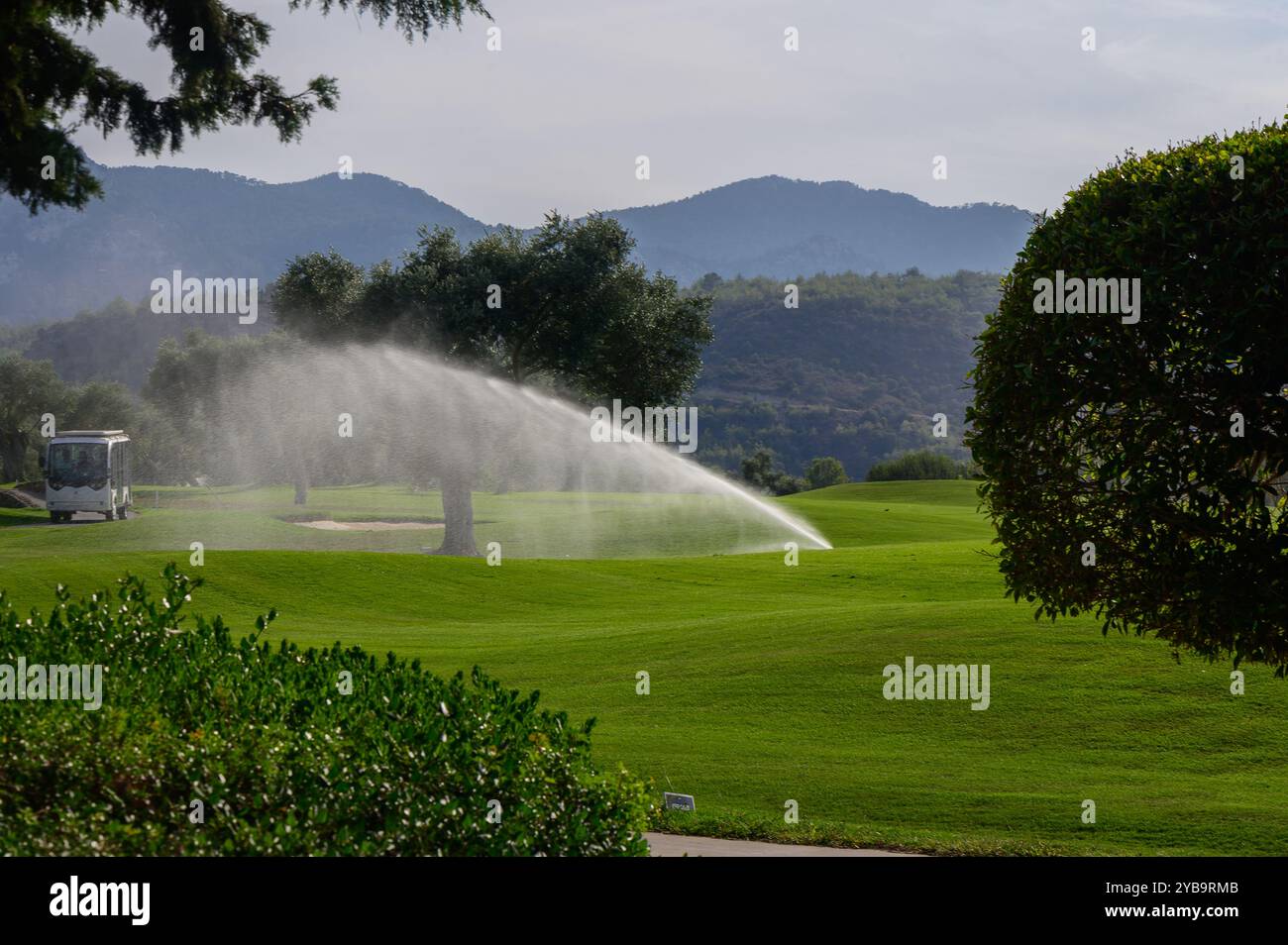 Un delicato getto di spruzzatori rivitalizzerà i verdi vibranti di un campo da golf in una mattinata di sole, con montagne maestose in lontananza che aumentano la t Foto Stock
