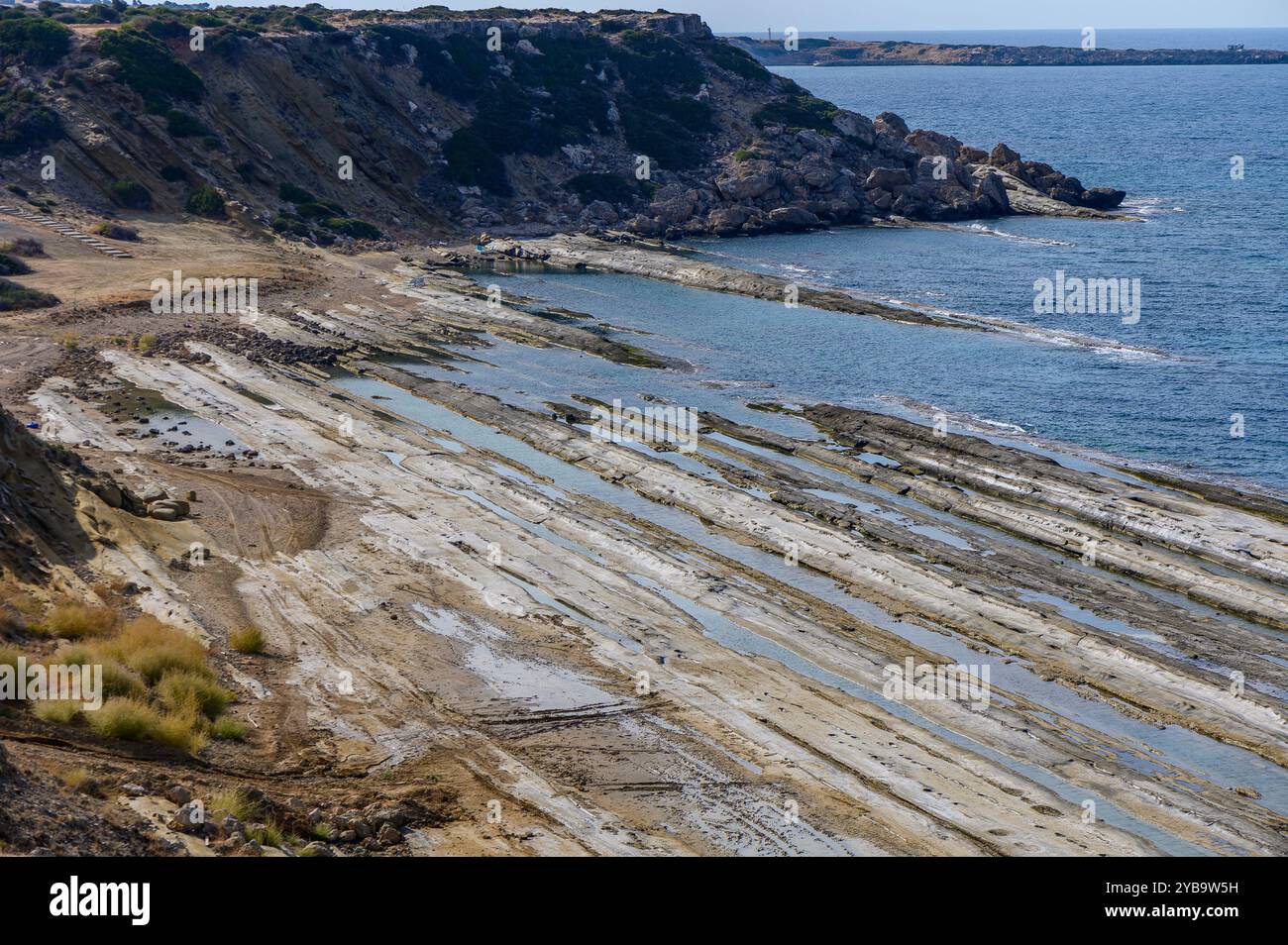 La costa rocciosa, scolpita dal tempo e dalla marea, rivela intricati motivi di terra umida e asciutta sotto il sole di mezzogiorno. Onde delicate si infrangono contro il Foto Stock