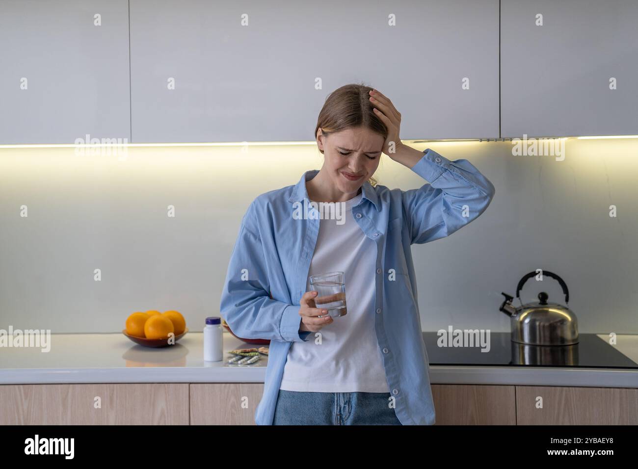 La donna depressa beve acqua dopo aver preso pillole per mal di testa, cercando di far fronte al dolore in cucina Foto Stock