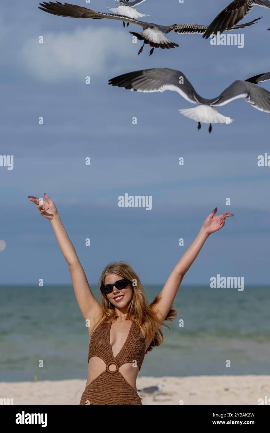 Una splendida donna nutre graziosamente i gabbiani su una spiaggia caraibica, circondata da acque turchesi e sabbia dorata, creando un pittoresco scenario di vacanza Foto Stock