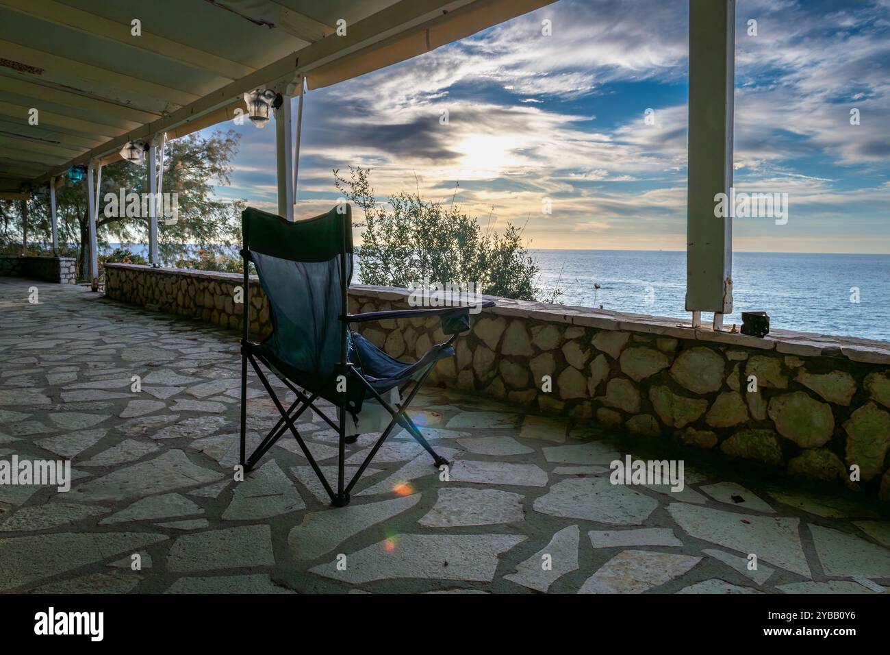 Una sedia pieghevole sul patio coperto di un edificio sul mare con una splendida vista del mare e delle nuvole del cielo del tramonto. Foto Stock
