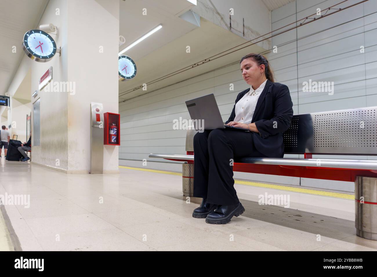 Giovane donna in costume da lavoro che lavora su un notebook mentre aspetta sulla piattaforma della stazione ferroviaria Foto Stock