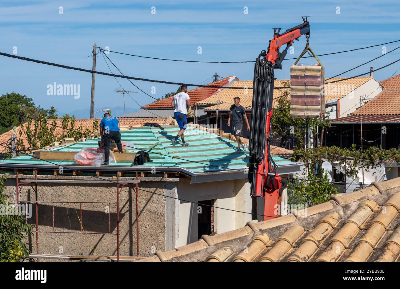 Lefkada, Grecia- 10.08.2024. Una gru di sollevamento che trasporta un fascio di tegole in ceramica al lavoratore che lavora sul tetto di un edificio in fase di ristrutturazione. Foto Stock