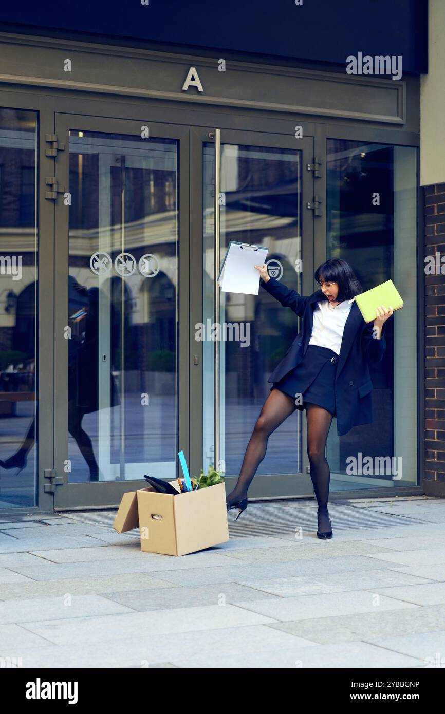 Giovane donna felice che balla allegramente dopo aver lasciato il lavoro, uscendo dall'edificio degli uffici con una scatola piena di cose Foto Stock