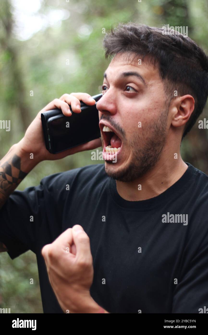 Un giovane frustrato urla nel suo cellulare durante un'intensa telefonata, in piedi in mezzo a una foresta pacifica Foto Stock