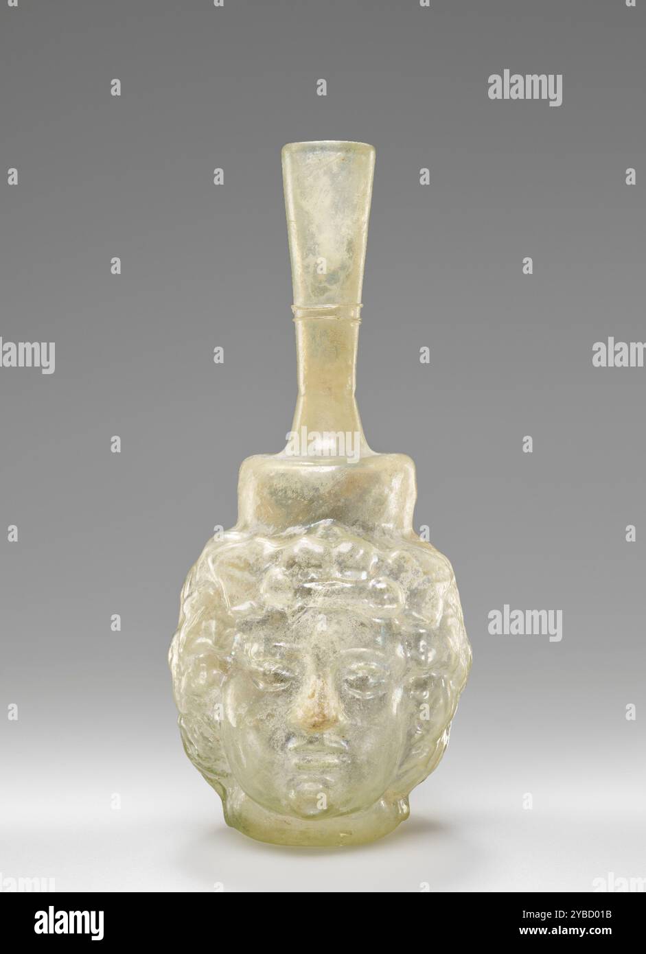 Head Flask, circa 2 ° secolo d.C. ulteriori informazioni: Questa fiasca con testa soffiata è stata realizzata in vetro incolore. Questo pallone è stato soffiato in due parti a forma di testa di un uomo (Dionysos) con una corona d'edera. Il corpo poggia su una base concava con un contrassegno. Piccole pareti cilindriche sorgono sopra la testa. Un collo svasato si solleva sopra la spalla appiattita. Il collo è decorato con due piste applicate al centro. Il pallone è in buone condizioni. La superficie presenta una leggera lucentezza iridescente. La punta del naso è stata ripristinata. Foto Stock