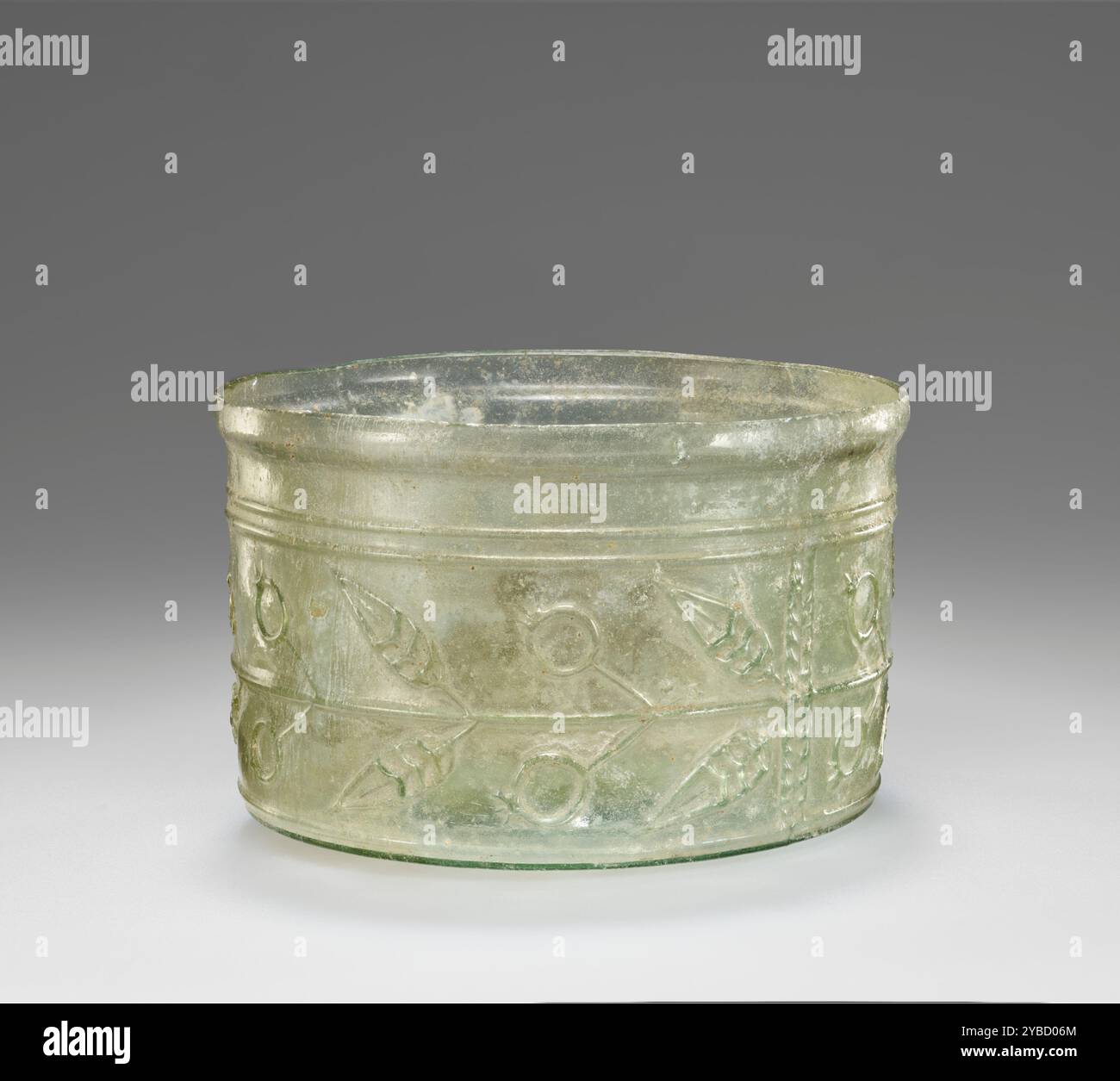 Coppa, i secolo d.C. informazioni aggiuntive: Tazza di vetro verde pallido gonfiata in uno stampo in tre parti indicato da cuciture che uniscono il corpo in due parti a una base. Corpo cilindrico decorato con melograno e fregio a foglia in rilievo su una base rialzata. Il cerchione è stato spaccato appena sopra un leggero trafilamento. Foto Stock
