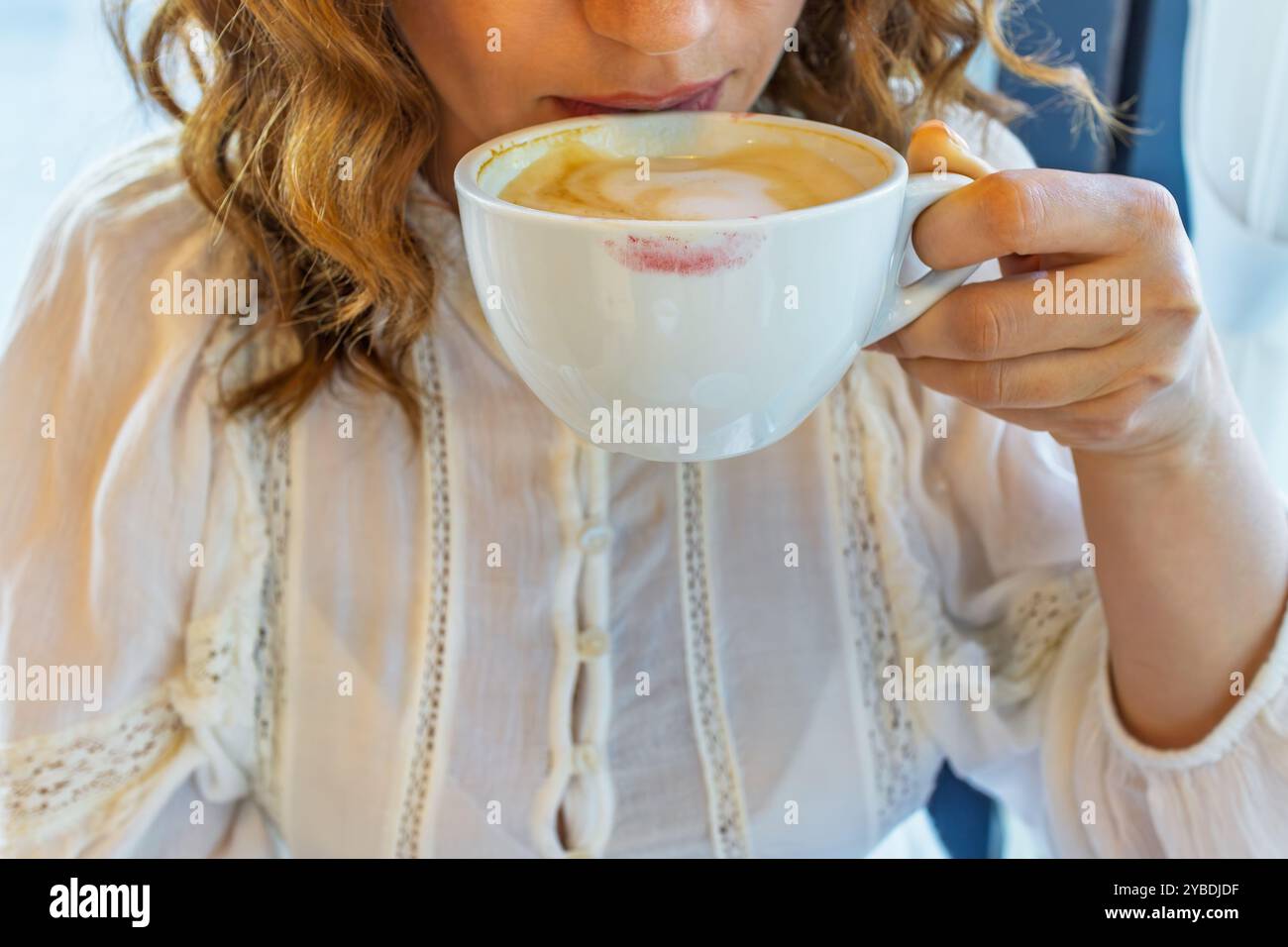 Sorseggia il caffè mattutino con colorante rossetto. Primo piano di una donna che sorseggia il caffè da una tazza bianca, lasciando un segno di rossetto. Il momento cattura un'atmosfera accogliente e accogliente Foto Stock