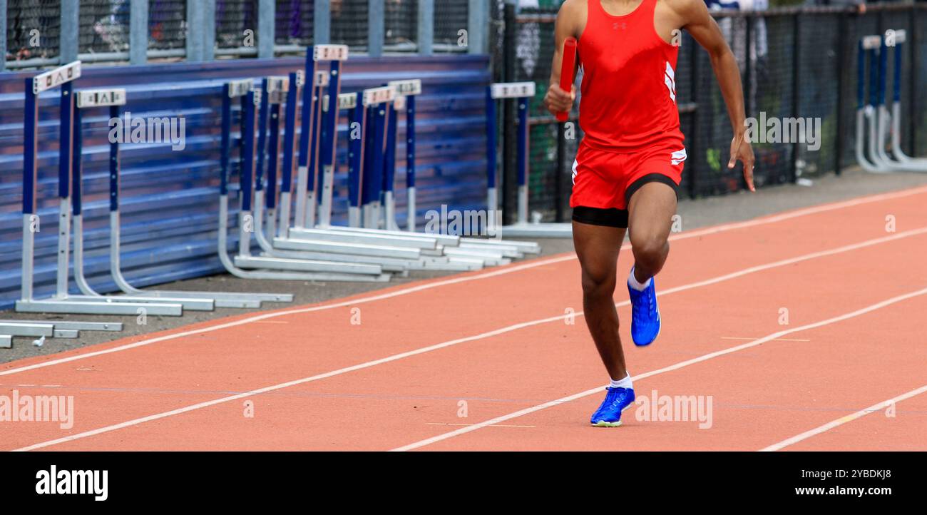 Vista frontale di un atleta afroamericano che corre in una gara di staffetta delle scuole superiori indossando un'uniforme rossa all'aperto. Foto Stock