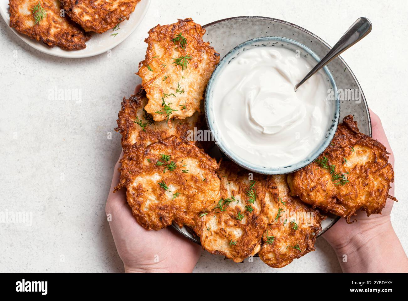 Mani che reggono piatto di patate fatte in casa o brocche di patate con panna acida, vista dall'alto. Latke o levivot ebraiche tradizionali. Foto Stock