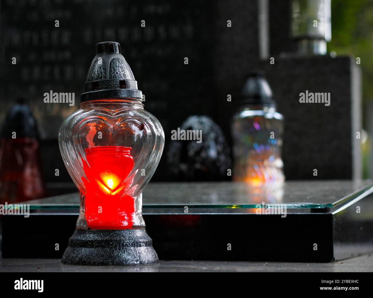 Primo piano di una candela rossa in fiamme in una lampada a forma di cuore del cimitero di vetro, altre candele del cimitero e una lapide con i nomi del defunto nella ba Foto Stock