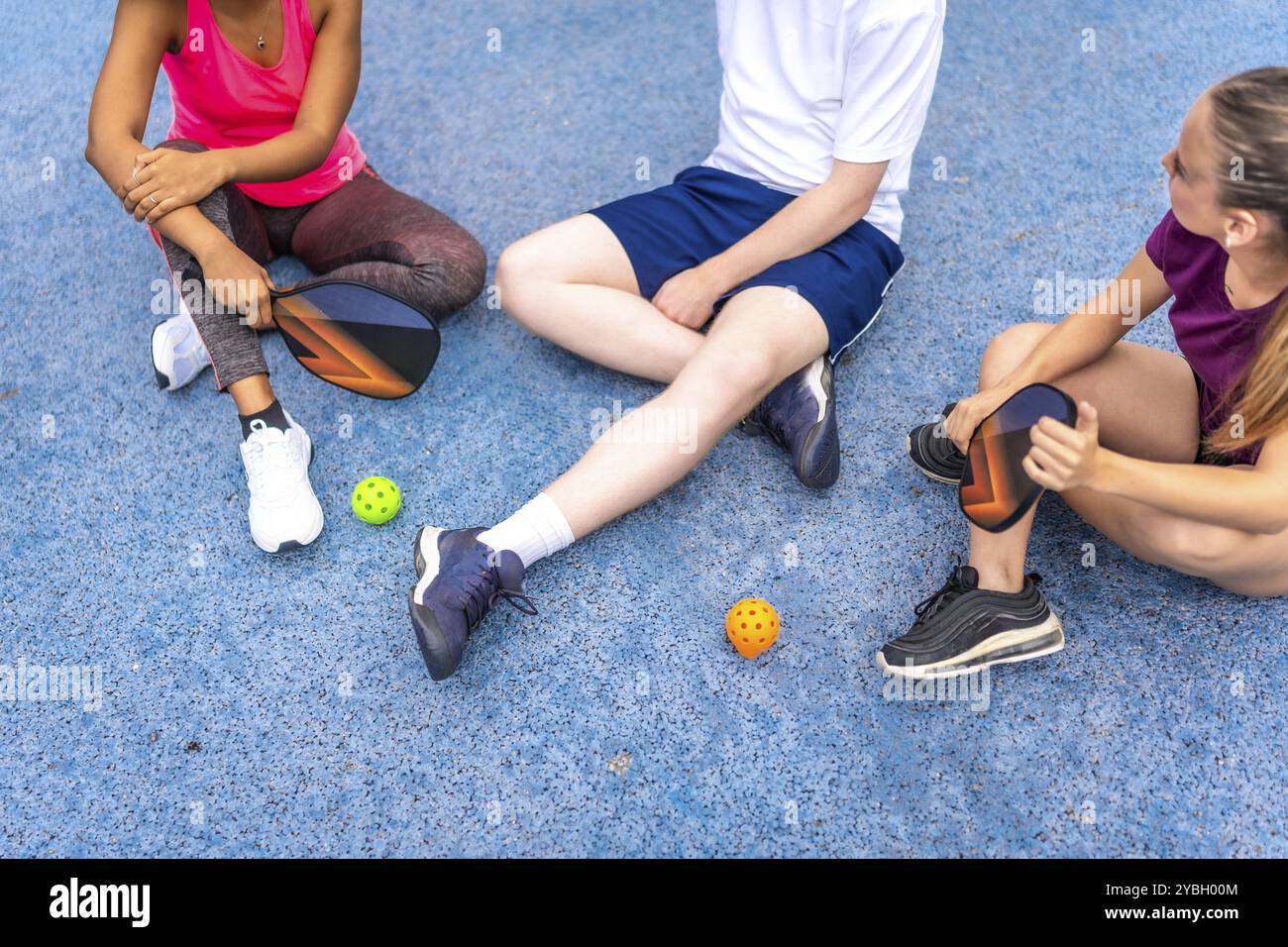 Foto ritagliata di un gruppo multietnico di amici seduti sul campo da baseball blu e chiacchierati Foto Stock