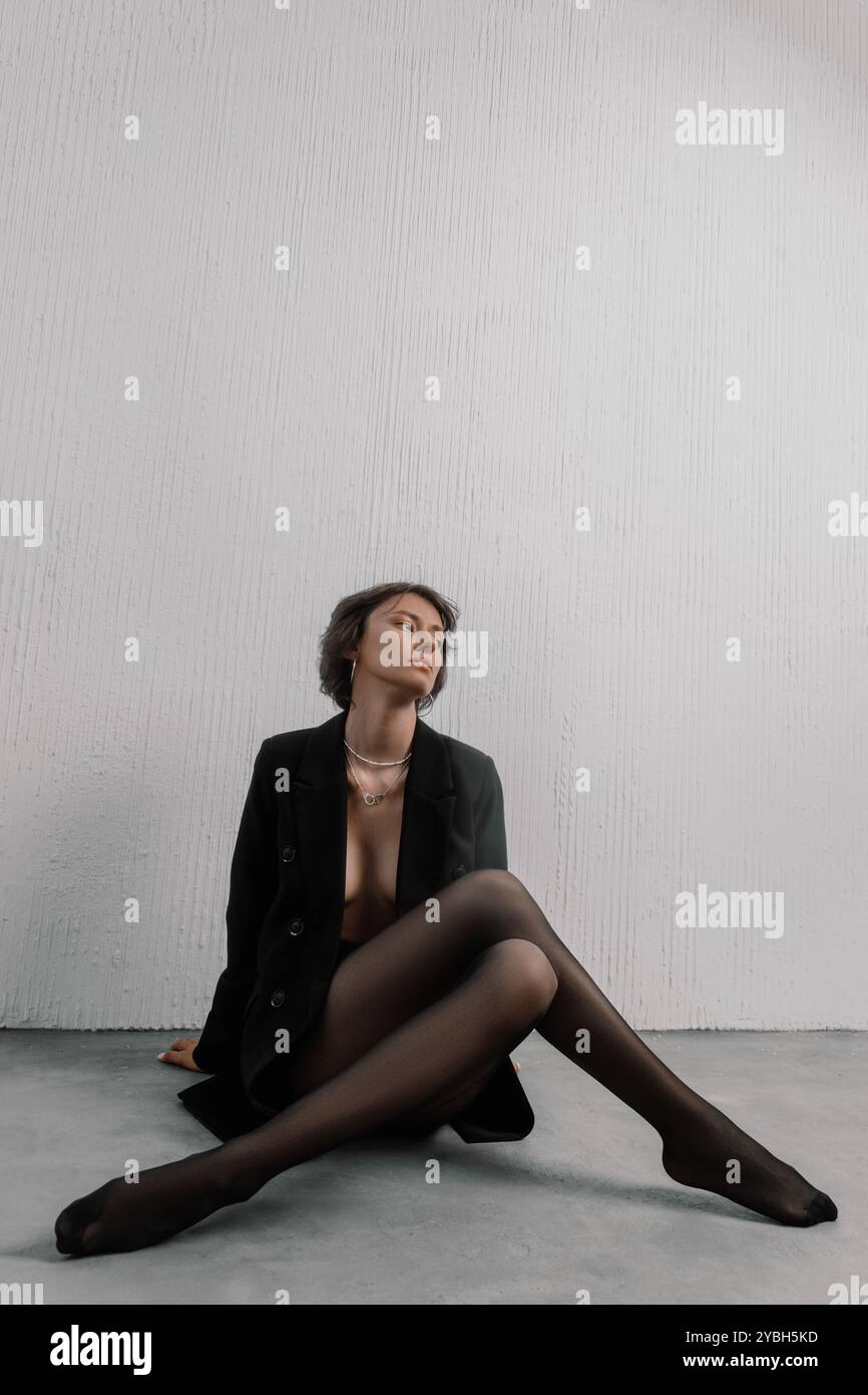 La donna siede graziosamente sul pavimento, indossando un blazer nero e calze. Guarda attentamente verso l'alto, circondata da un colore chiaro e pulito Foto Stock