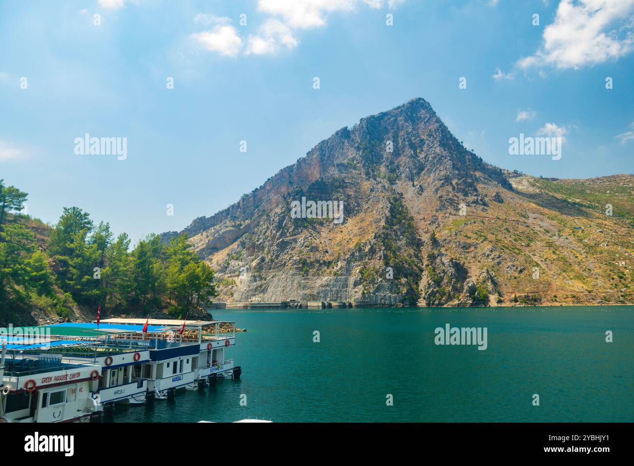 Vista panoramica della diga di Oymapinar in lontananza contro un cielo blu e un bacino idrico verde e barche in primo piano in una soleggiata giornata estiva Foto Stock