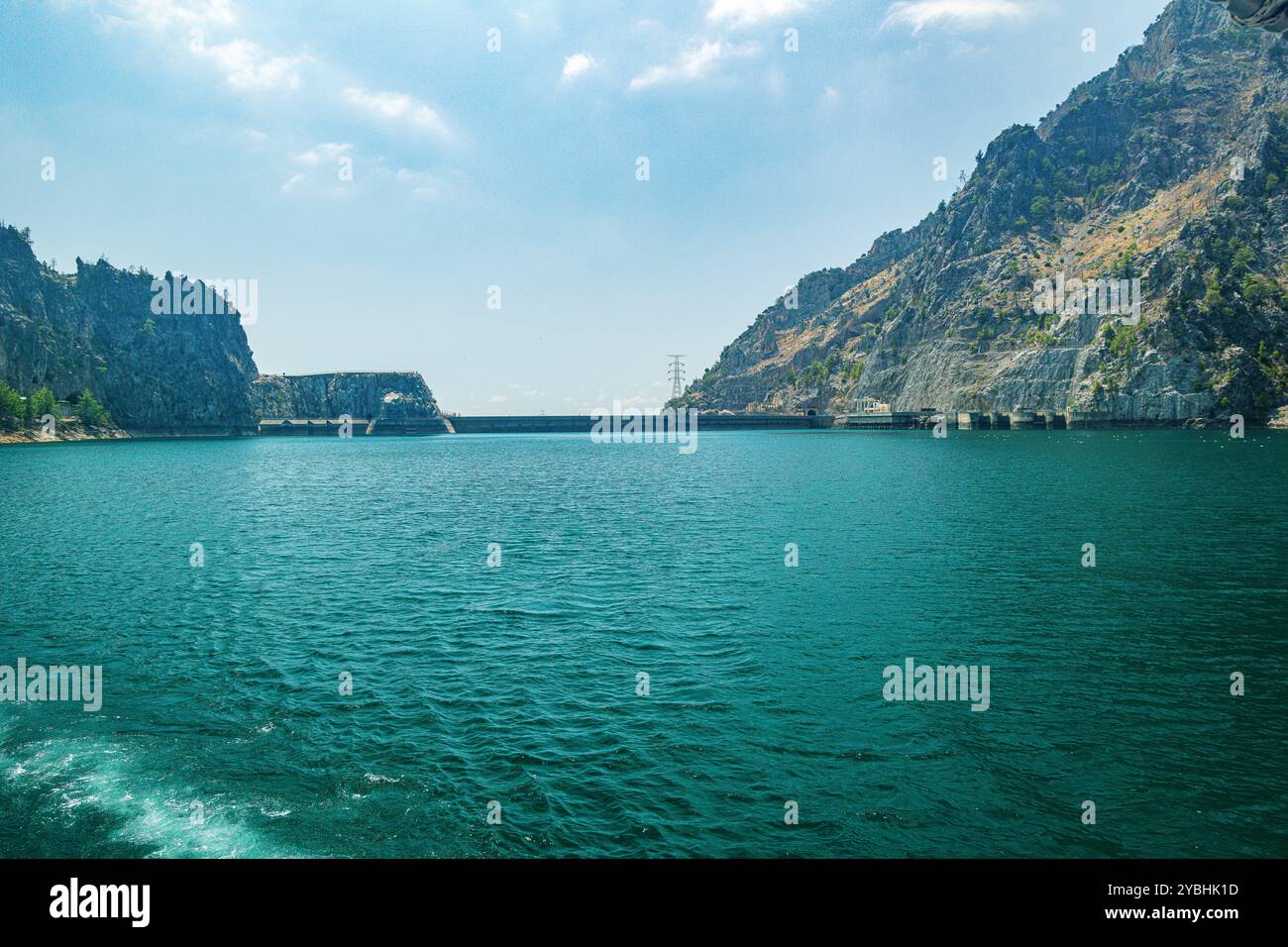 Vista panoramica della diga di Oymapinar in lontananza contro un cielo blu e un bacino idrico verde in primo piano in una soleggiata giornata estiva Foto Stock