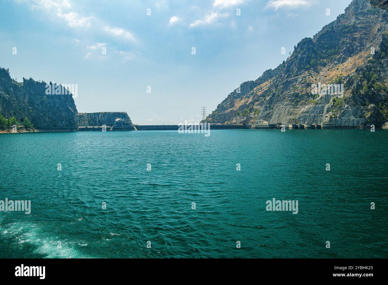 Vista panoramica della diga di Oymapinar in lontananza contro un cielo blu e un bacino idrico verde in primo piano in una soleggiata giornata estiva Foto Stock