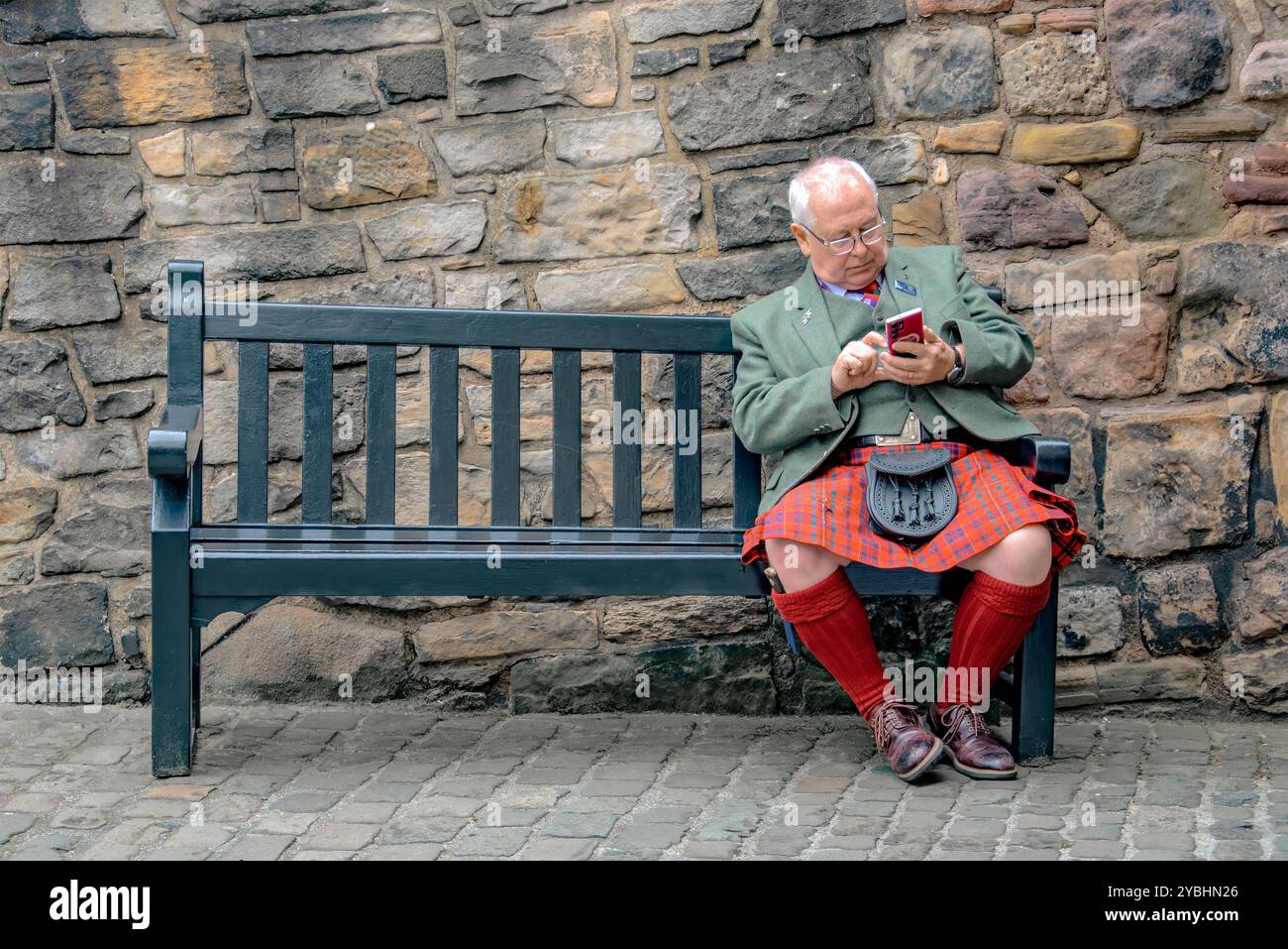 Castello di Edimburgo, Scozia, 5 maggio 2024: Uno scozzese, vestito con uniformi tradizionali di kilt e sporrans, si rilassa su una panchina e guarda il suo cellulare. Foto Stock