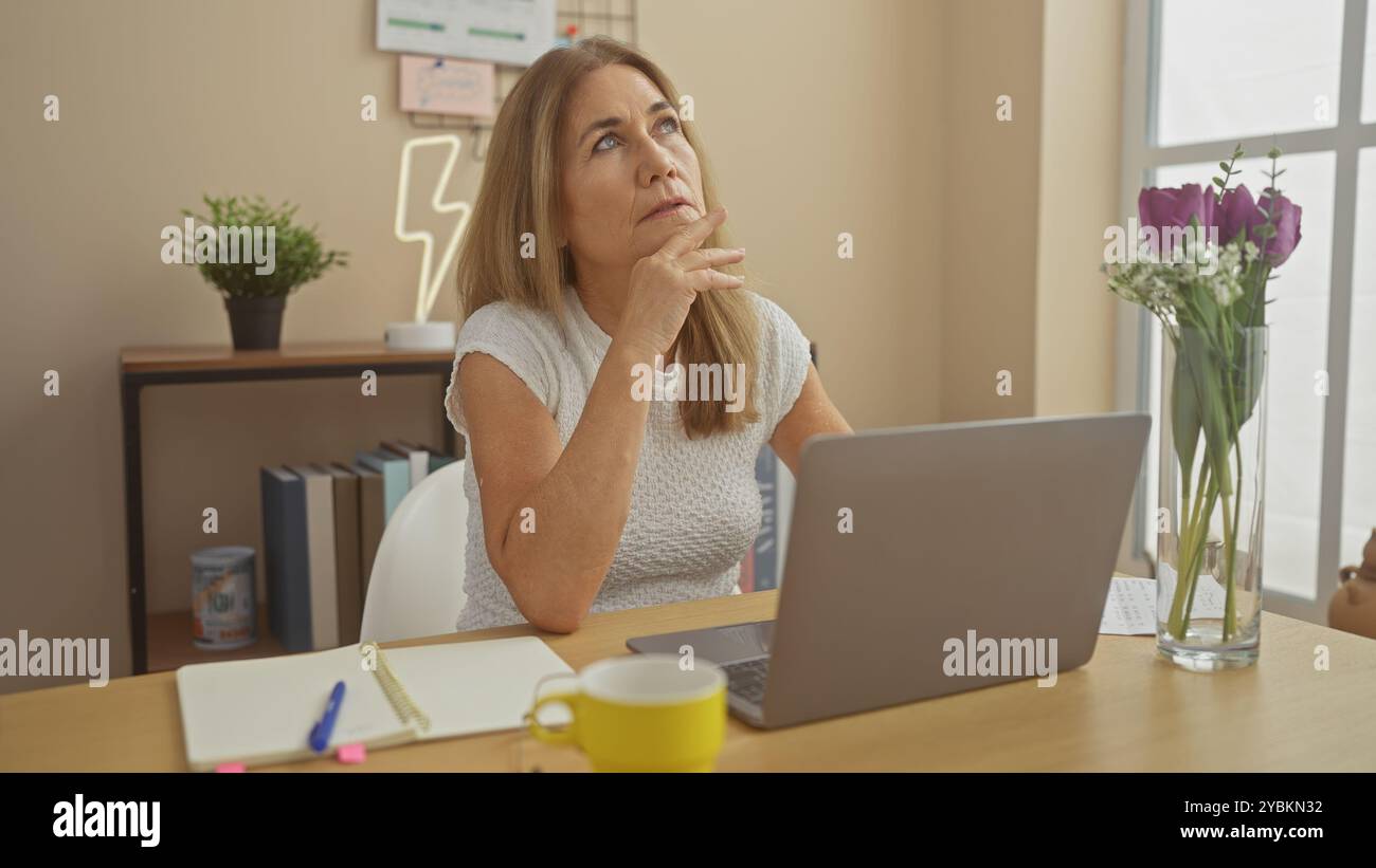 Donna matura che contempla mentre lavora con un notebook in un ufficio domestico con fiori e tazza da caffè. Foto Stock