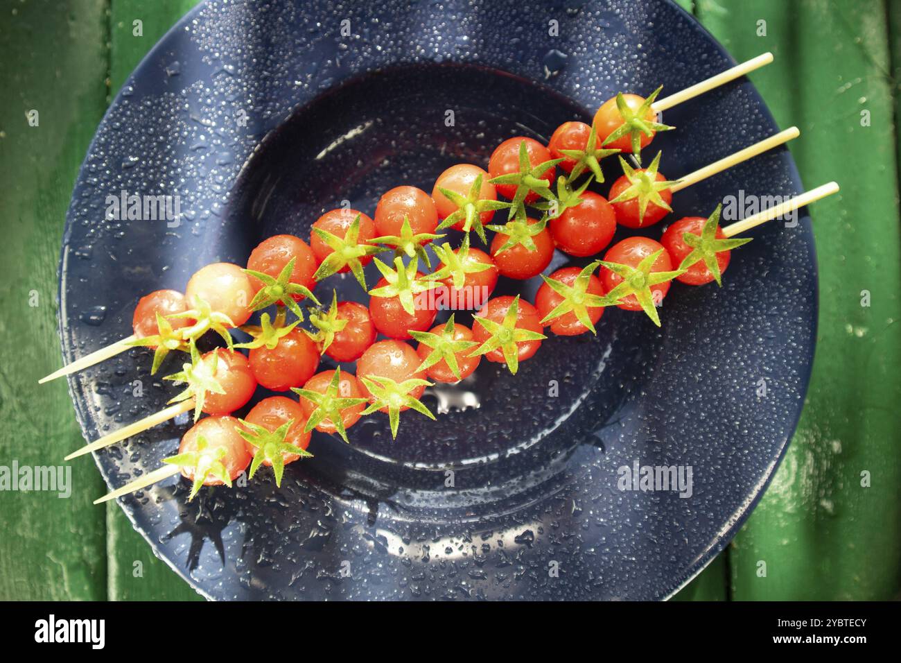 Rappresentazione fotografica di tre spiedini realizzati con pomodori ciliegini rossi e gustosi di Pachino, Sicilia Italia, posti su un piatto blu Foto Stock