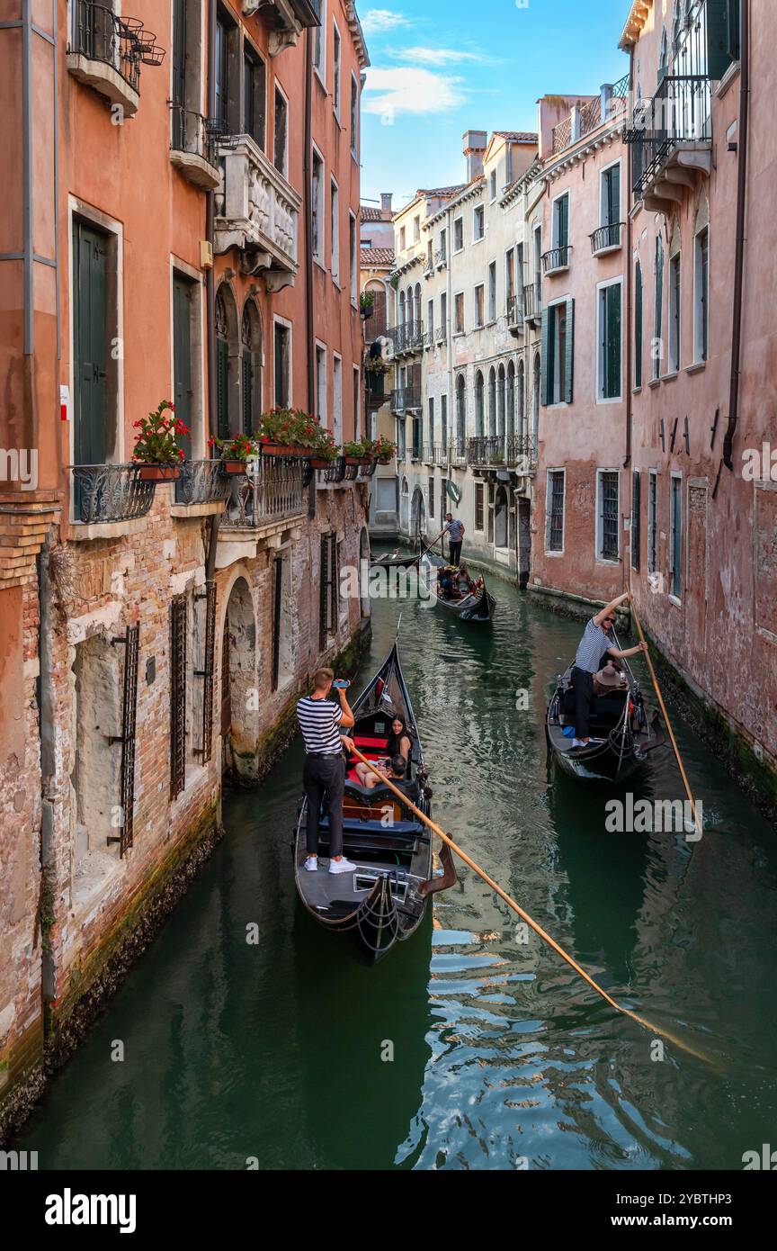 Dall'alto di anonimi gondolieri maschi che guardano in basso mentre remi in piedi su lunghe barche strette nell'acqua del canale di Venezia tra gli edifici in pieno giorno Foto Stock