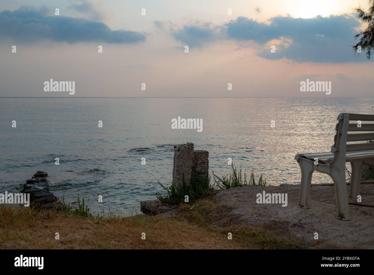 Una panchina bianca sotto l'ombra di un albero sulla spiaggia di Zante per rilassarsi in tranquillità durante l'ora d'oro. Un posto tranquillo vicino al mare. Foto Stock