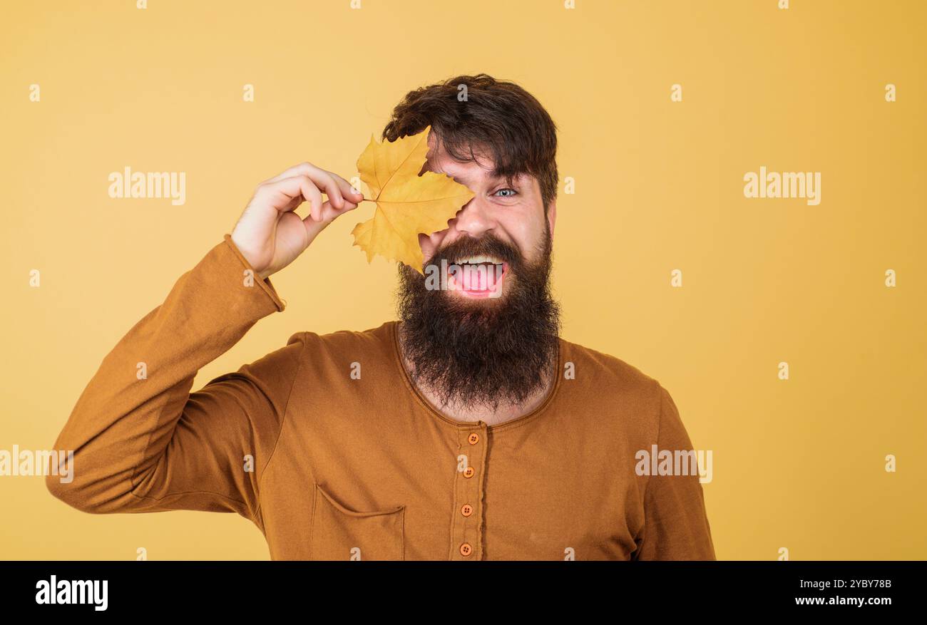 Modello maschile autunnale. Uomo con barba felice che copre l'occhio con foglie d'acero autunnali. Moda autunnale stagionale. Vestiti autunnali per uomini. Bell'uomo in un elegante orang Foto Stock