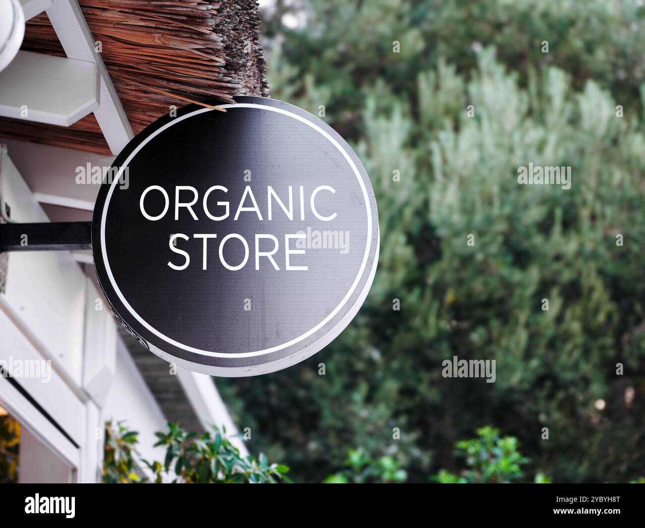 Un cartello nero rotondo con lettere bianche che recitano il negozio organico è appeso all'esterno di un edificio con alberi verdi sullo sfondo. Foto Stock